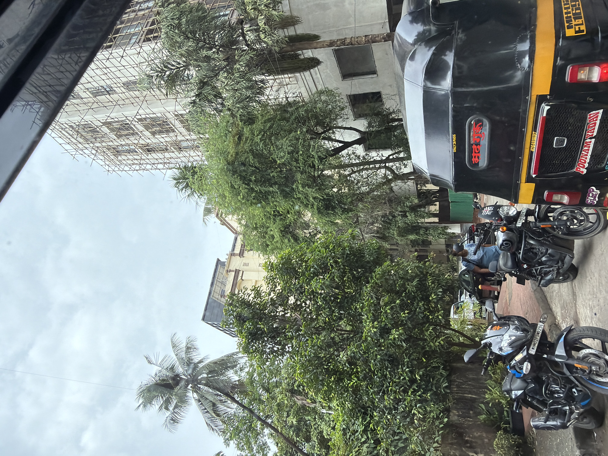 The image depicts a street scene in Mumbai, India, with an overcast sky and lush greenery. In the foreground, two black motorcycles are parked, one in sharper focus than the other. The motorcycle on the left has blue accents and a license plate that reads "MH 02FS 0296". To the right of the motorcycles is the rear of a black auto-rickshaw with yellow accents. It displays text in red and yellow on its back, including what appears to be a name "POONAM YADAV". Behind the vehicles, a woman wearing a blue top is visible.
In the background, a multi-story building is under construction, covered in scaffolding. Adjacent to this building is another older, cream-colored building with arched windows. Tall palm trees and other dense green foliage are scattered throughout the scene, suggesting a tropical urban environment. Several cars are parked further down the street. The overall impression is a typical street view in a bustling Indian city, with elements of both ongoing development and established urban life.