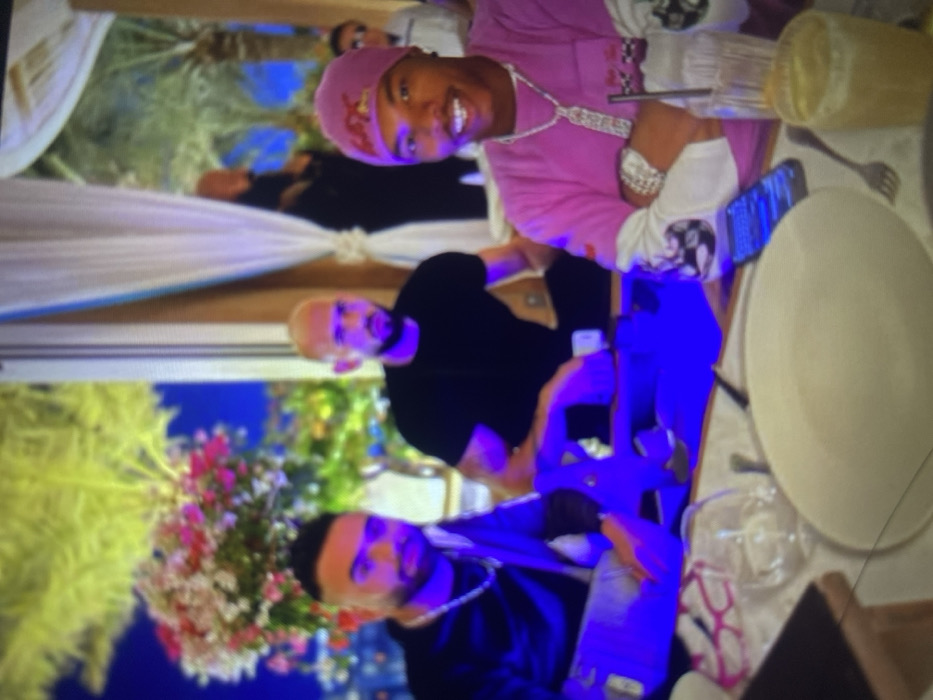Three men are seated at a table. The man at the top wears a pink outfit with a matching head covering. Below him is a man in a black shirt. The bottom man wears a dark jacket with a silver chain. A white plate, silverware, and drinking glasses are arranged on the table. The scene is brightly lit, possibly outdoors. There are white draperies and a floral arrangement behind the men. Two phones appear to be on the table.