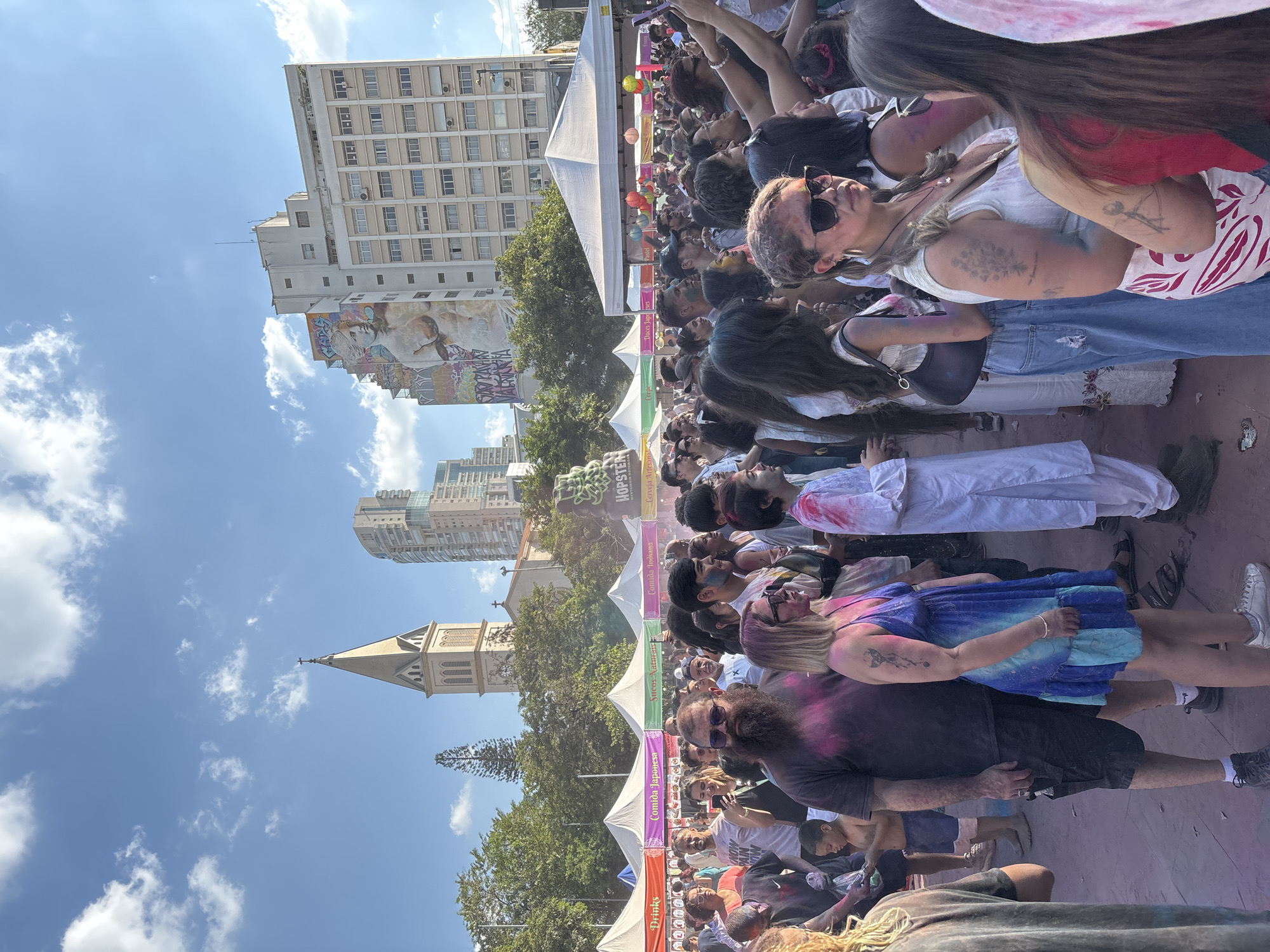 The image captures a crowded outdoor event, possibly a festival or celebration, taking place in a public space in São Paulo, Brazil. The crowd is dense with people of diverse ages and appearances. Many individuals are covered in colored powder, indicating participation in a Holi-like or similar color-based festival. A series of white vendor tents lines one side of the scene, each with signage indicating the type of food or product being sold.
In the background, two prominent buildings are visible: a tall, multi-story building with a mural on its side and a church with a steeple. The sky above is bright blue with scattered clouds. The overall environment suggests a warm, sunny day in an urban setting. The location can be identified as the area around the São Paulo Cathedral, located in the city's central area.