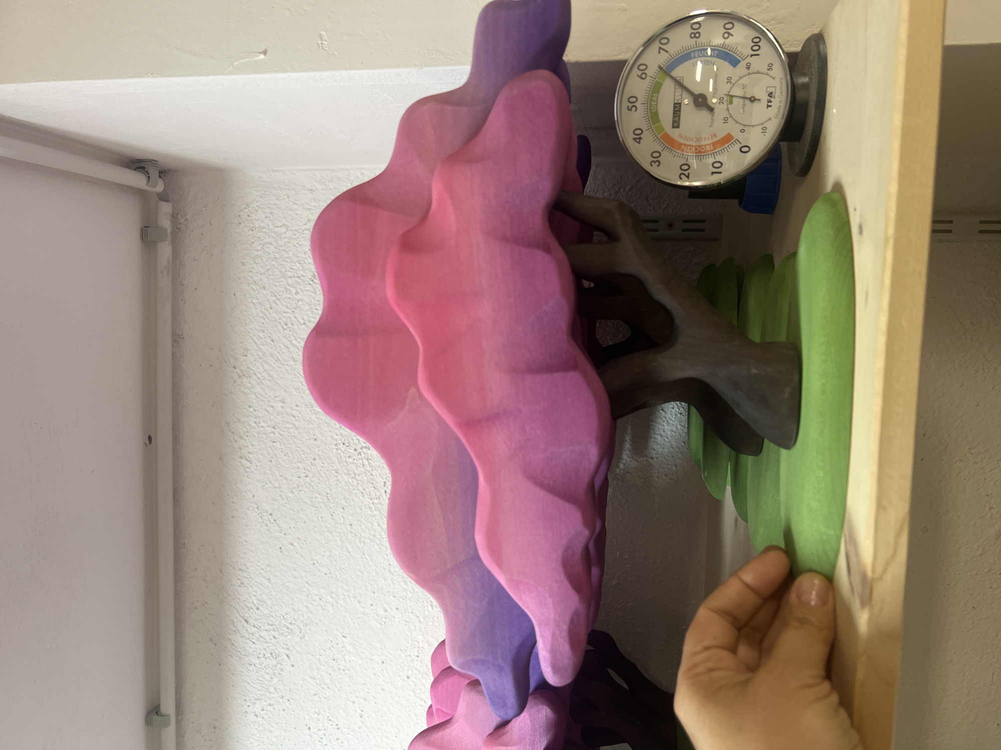 The image shows a close-up of a wooden decorative tree with pink and purple clouds as foliage. The tree is placed on a green, grassy base, and is situated on a wooden shelf. To the right of the tree is a round analog hygrometer and thermometer. A human hand is visible in the lower left corner, gently touching the green base of the tree. The background consists of a textured white wall and some white pipes affixed to the wall and ceiling. The scene appears to be indoors, possibly in a workshop or storage area.