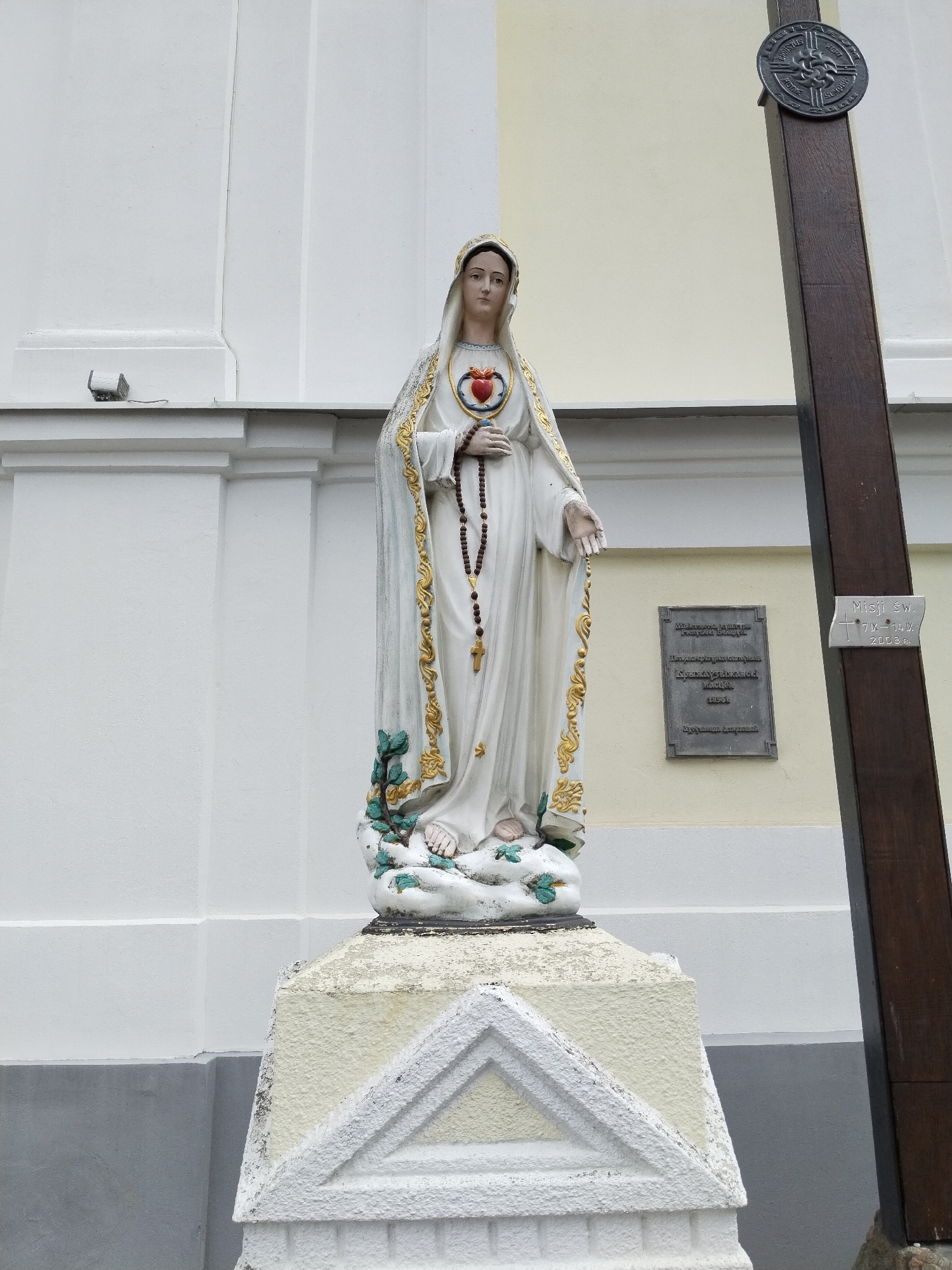 A full-body statue of the Virgin Mary stands on a pedestal outside a building in Brest, Belarus. The statue depicts Mary with a halo and a heart pierced by a sword, symbolizing her immaculate heart. She wears a white robe with gold trim and holds a rosary. The pedestal is decorated with a triangular motif. To the right of the statue, a wooden cross with a circular emblem is positioned. A plaque on the wall behind the statue provides historical information about the location. The building appears to be a church or religious institution, with a white facade and a light yellow accent wall. The sky is overcast. The image is taken from a low angle, looking up at the statue.