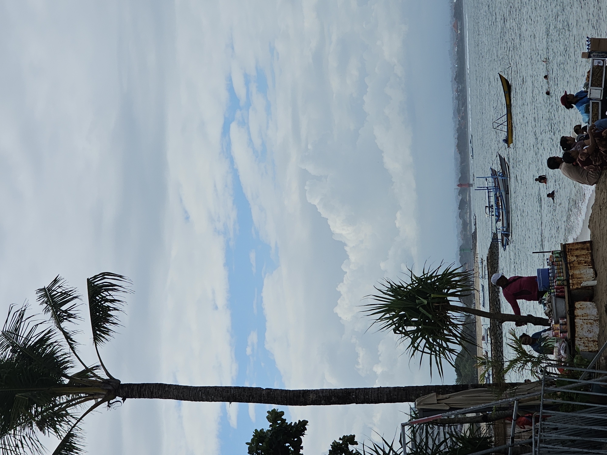 An outdoor, slightly overcast scene depicts a beach in Denpasar, Indonesia. In the foreground, a tall palm tree stands to the left. Below it, a small vendor stand is set up on the sand, with two vendors and several customers. The vendors are attending to a display of bottled drinks and packaged snacks. Customers are seated nearby, observing the wares.
In the middle ground, several boats are anchored or moving on the water. The sea extends to the horizon, where a distant coastline is visible with some structures and foliage. The sky is filled with a mixture of white and grey clouds, with patches of blue visible. The overall impression is a casual beach day with local commerce taking place.