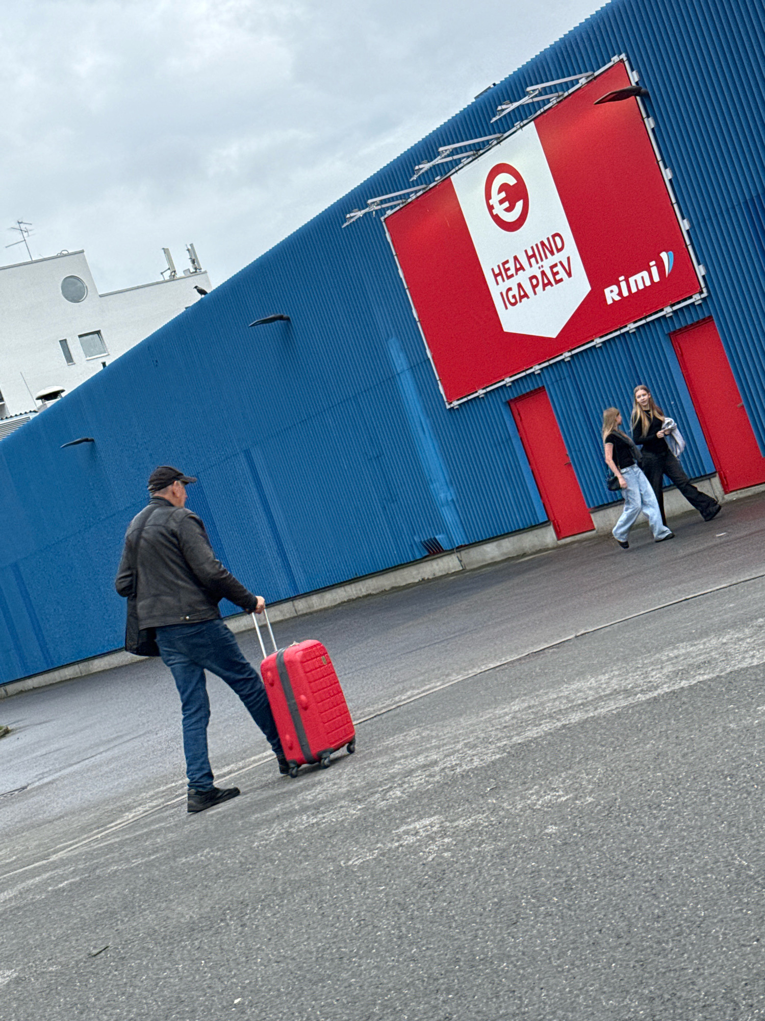The image depicts an outdoor scene in Tallinn, Estonia, in front of a large, blue corrugated metal building with a prominent red sign at the top. The sign features a white section with a euro symbol and text in Estonian that reads "HEA HIND IGA PÄEV" (Good Price Every Day), and a smaller section with the "Rimi" logo. Two red doors are visible on the building's facade.
In the foreground, a man wearing a black cap, black leather jacket, and blue jeans is pulling a bright red suitcase on wheels across an asphalt surface. To the right of the man, two young women are walking away from the camera. One woman is wearing a black top and light blue wide-leg jeans, and the other is wearing a black top and black flared pants, carrying a bag over her shoulder. The ground is wet, suggesting recent rain. In the background, a white building with a circular window and antenna is partially visible. The sky is overcast.