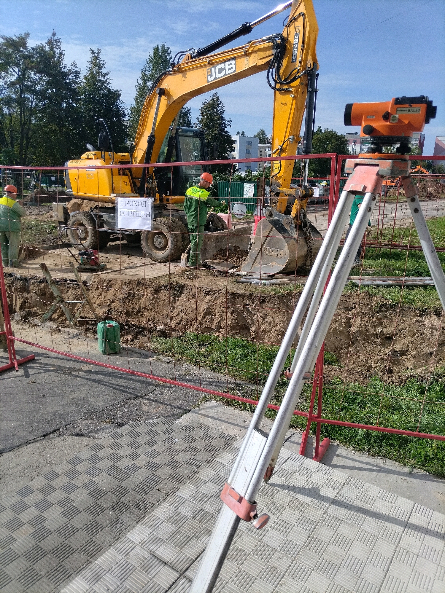 The image shows a construction site in Hrodna, Belarus, with an excavator and two construction workers. The excavator is a yellow JCB model. One worker, wearing a green uniform and an orange helmet, is standing near the excavator's bucket. The other worker, also in a green uniform and orange helmet, is standing on the other side of the excavation pit. A sign in Russian is attached to the excavator, reading "ПРОХОД ЗАПРЕЩЕН" (Passage Forbidden). The excavation pit is fenced off with red barriers and wire mesh. In the foreground, a surveying tripod with an orange leveling instrument is set up. The ground surface consists of gray patterned tiles and asphalt. Trees and buildings are visible in the background. The sky is blue with some clouds.
