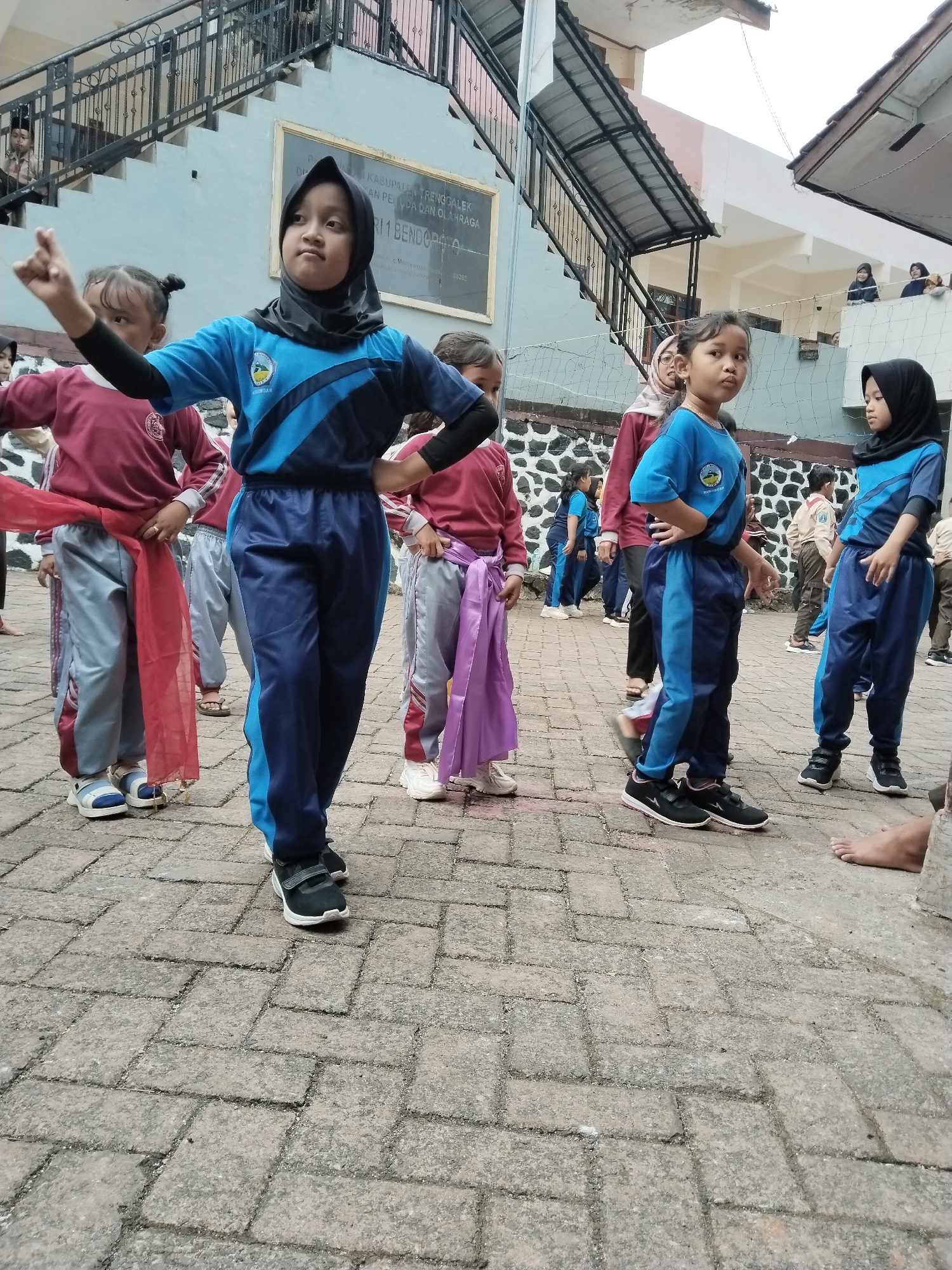 In Bendoroto, Indonesia, children are gathered outdoors on a brick-paved area, possibly for a school event or cultural performance. In the foreground, a young girl wearing a black hijab and a blue and black tracksuit is striking a pose with her right hand raised, index finger pointing. To her left, another girl in a similar tracksuit is partially visible. Several other children are present, some wearing maroon and gray tracksuits with red and purple sashes, others in scout uniforms. Behind the children, a multi-story building with an outdoor staircase and a metal railing is visible. A sign on the building reads "KABUPATEN TRENGGALEK DINAS PEMUDA DAN OLAHRAGA RT 1 BENDOROTO". The sky is overcast, suggesting a cloudy day.