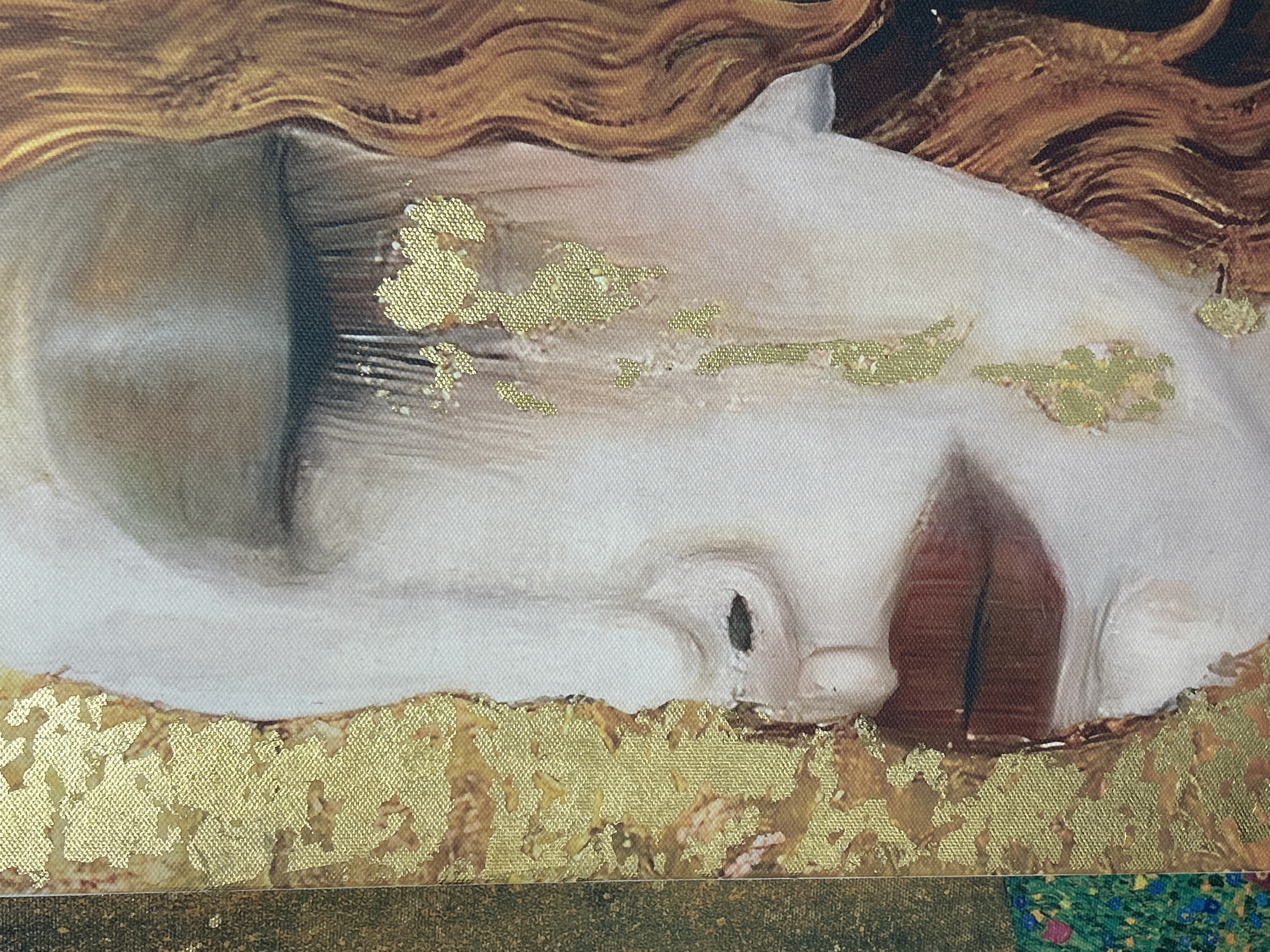 This close-up image displays a section of an artwork, likely a painting or print on canvas, featuring a serene female face in profile. The face is rendered in pale, smooth tones, with closed eyes, a delicate nose, and full, reddish-brown lips that are slightly parted. Flowing golden-brown hair cascades around the head, painted with wavy textures.
A significant element of the artwork is the extensive use of gold leaf or highly textured gold paint. These shimmering, golden applications appear as scattered, irregular patches over the face, particularly near the eye and cheek, creating a decorative, almost ethereal effect. Larger, more continuous bands of gold are visible beneath the face and along the bottom edge of the primary subject, suggesting an ornate background or an integral part of the composition. The gold often shows a distinct, patterned texture, highlighting the underlying canvas weave.
The artistic style, with its combination of figurative representation and rich gold embellishments, evokes a sense of Symbolism or Art Nouveau, reminiscent of artists like Gustav Klimt. The visible canvas texture throughout the image underscores the physical nature of the artwork. At the very bottom of the frame, a narrow band of dark material gives way to a hint of a vibrant, textured pattern in greens, blues, and pinks, possibly another decorative element within the artwork or a surrounding frame. There is no visible text in the image.