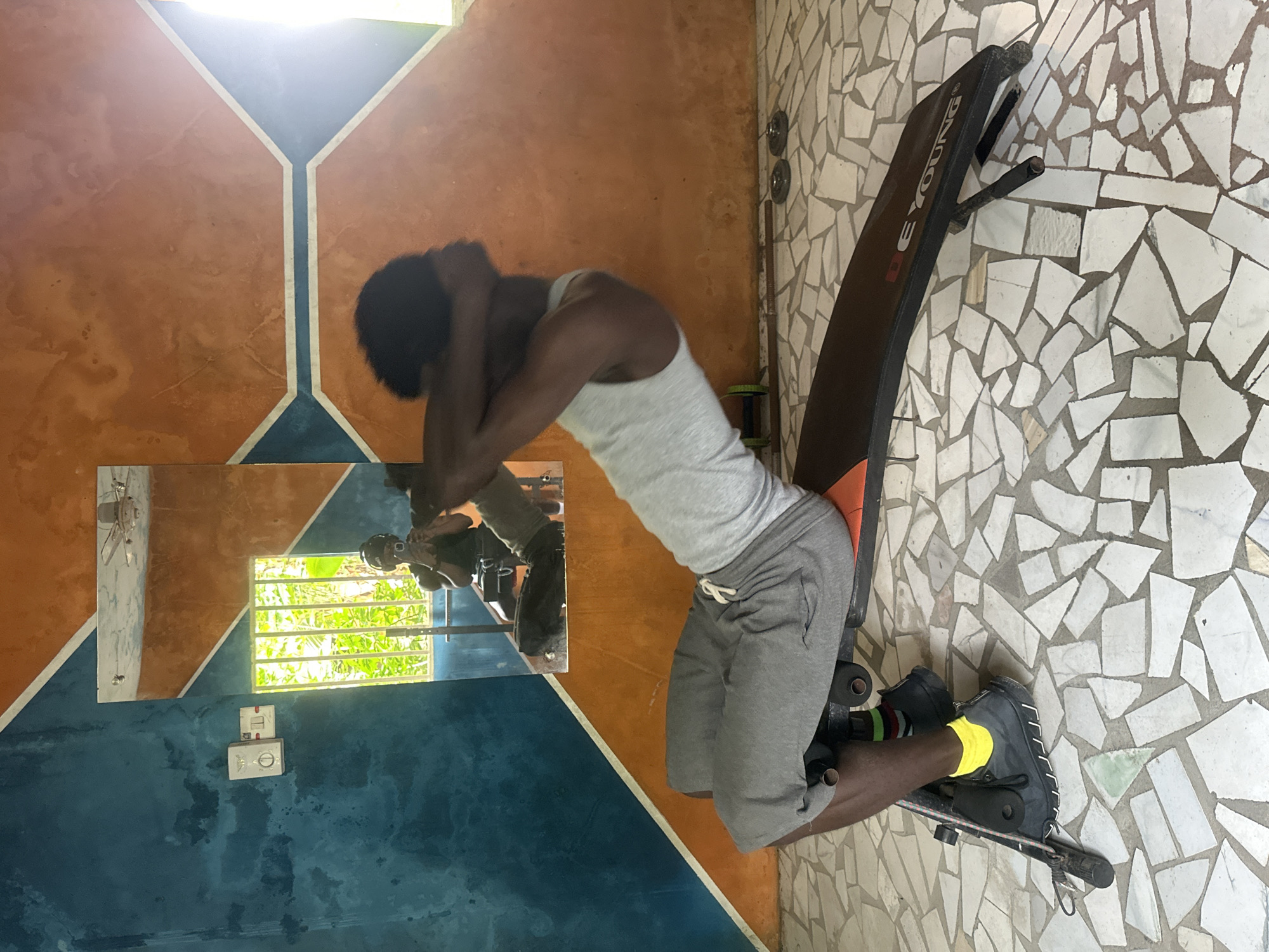 In Badagry, Nigeria, a Black male is performing sit-ups on an exercise bench. He is wearing a grey tank top, grey shorts, black shoes, yellow socks, and striped socks. His hands are clasped behind his head. The exercise bench is positioned on a floor tiled with irregular pieces of white, grey, and green material. The background features a wall painted with a diagonal geometric design in blue and orange. A mirror on the wall reflects the man, a window with green foliage outside, a ceiling fan, and a light switch. Dumbbells are visible on the floor near the bench.