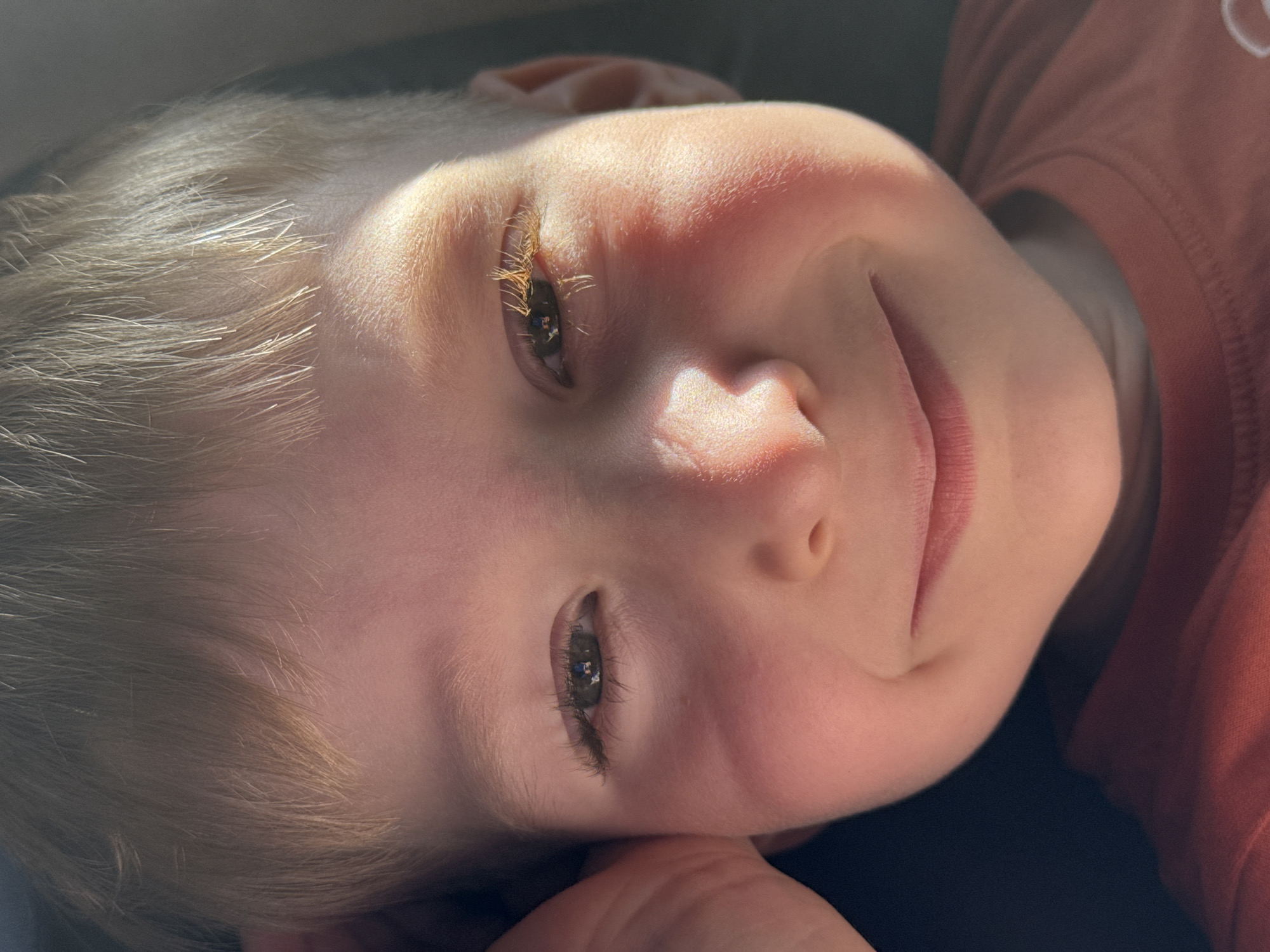 In Vykhodnoi, Russia, a close-up, eye-level shot captures a young boy with fair hair and fair skin. The boy is looking directly at the camera, his head tilted slightly and his left hand resting on his cheek. His eyes are open, with the right eye appearing more prominent due to lighting. The boy's lips are parted in a gentle smile. A strand of hair falls across his forehead, and sunlight illuminates his face, highlighting his features and casting shadows. He is wearing a rust-colored t-shirt. The background is dark and indistinct, suggesting an indoor or shaded outdoor setting.