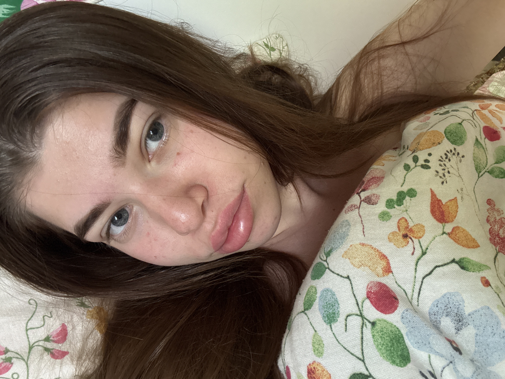 A young woman with fair skin, blue eyes, and brown hair is lying down, possibly in a bed. She is positioned at an angle, looking towards the camera with a slightly downward gaze. Her hair is loose and spread around her head. She appears to be resting on a pillow or blanket with a floral pattern in various colors including orange, red, green, and blue. The background is indistinct and out of focus. There is no information to suggest the location is in Мещёры, Russia or anywhere else.