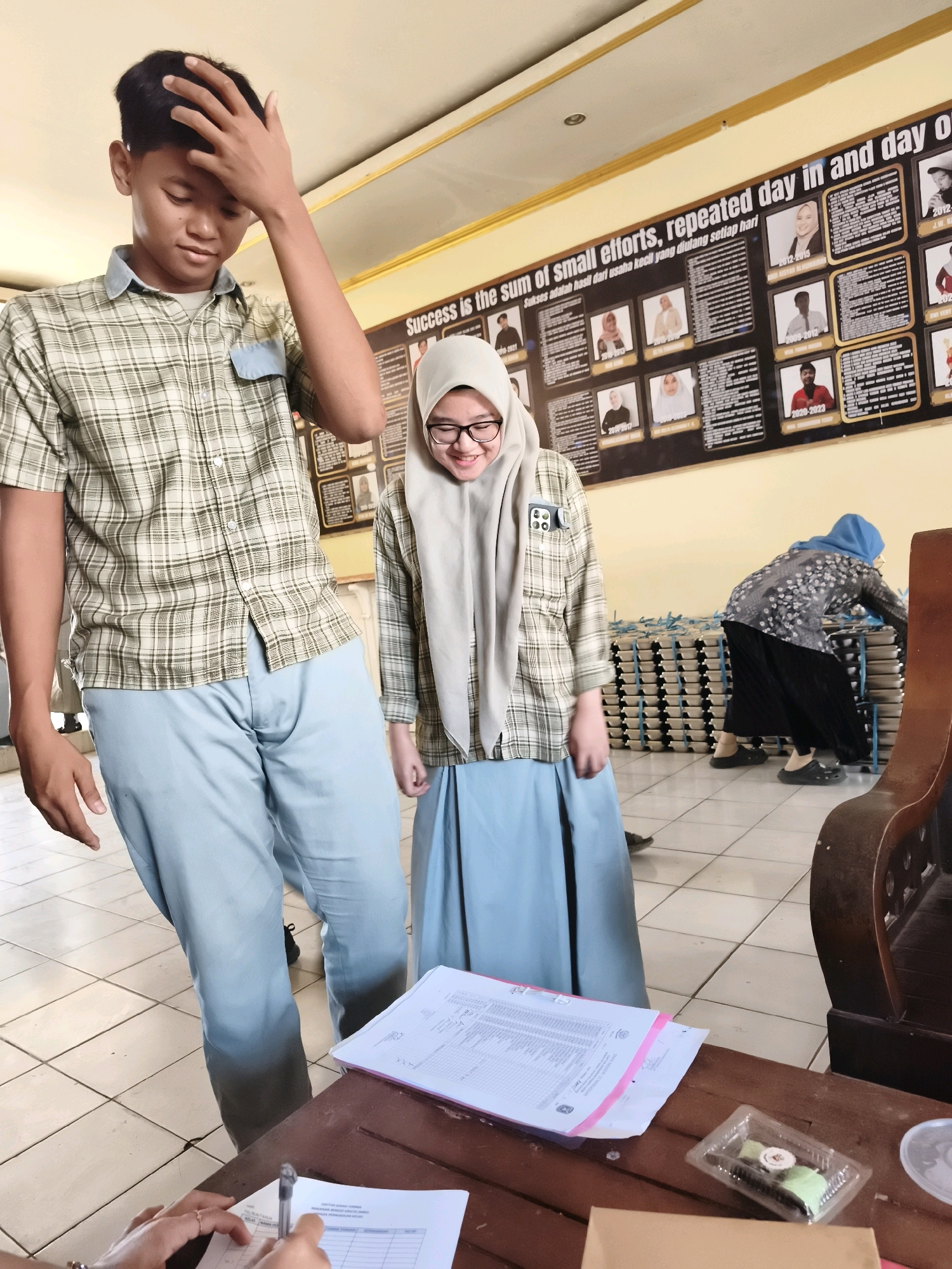 In a room with beige walls and a yellow ceiling, a young man stands with his hand on his head, looking down. He wears a plaid shirt and light blue pants. To his right, a young woman in a hijab and glasses smiles, also wearing a plaid shirt and a light blue skirt. Behind them, a bulletin board displays photos and text in English and Indonesian. In the background, a woman in a blue hijab and a patterned shirt bends over a stack of trays. In the foreground, a wooden table has papers, a pen, and a small container of food. The floor is tiled. The setting appears to be a school or community center in Makassar, Indonesia.