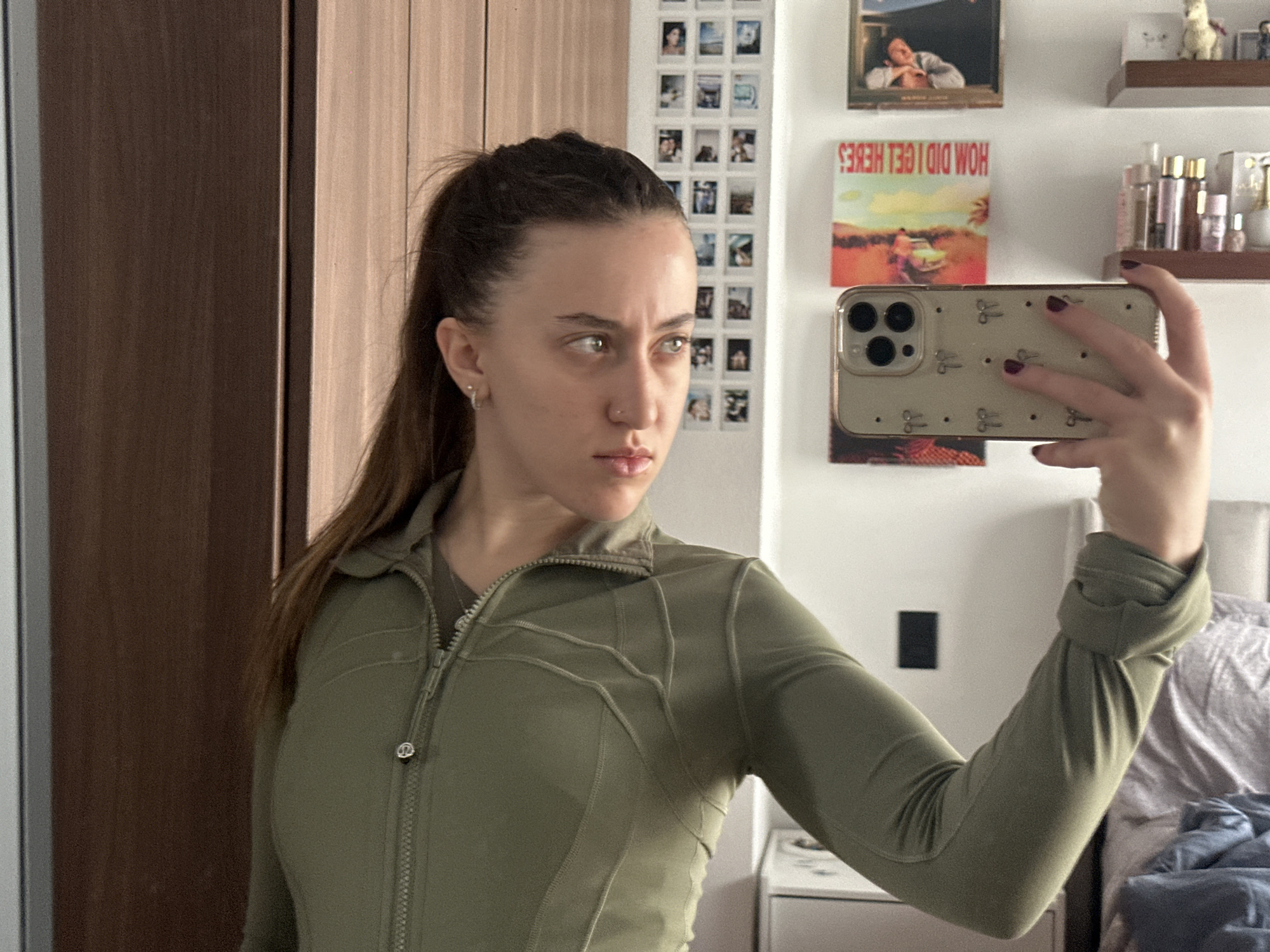 A young woman is captured in a mirror selfie, standing in what appears to be a personal room in Huixquilucan de Degollado, Mexico. She has long, dark brown hair styled in a high ponytail and is wearing an olive green, long-sleeved athletic jacket, possibly from Lululemon, identifiable by the distinctive logo on its zipper pull. She has small hoop earrings and a nose piercing. Her expression is serious and focused as she looks to her left, slightly away from the camera.
She holds a light-colored smartphone, likely an iPhone given its triple-camera system, enclosed in a beige or light gold case adorned with small, repetitive scissor-shaped motifs and dark dots. Her fingernails are painted a dark red or burgundy color.
The background, visible in the mirror's reflection, reveals a domestic setting. On the left, a dark brown wooden door or wardrobe is seen. The white wall on the right is decorated with a grid of numerous small, square polaroid-style photographs. Above her phone, a framed picture is displayed, and below it, an album cover or poster features the reversed text "HOW DID I GET HERE." Higher up on the wall, a small wooden shelf holds various bottles and a white llama figurine. Part of a bed with grey and blue bedding is visible in the lower right, suggesting the selfie was taken in a bedroom during daytime, indicated by the bright, even lighting.