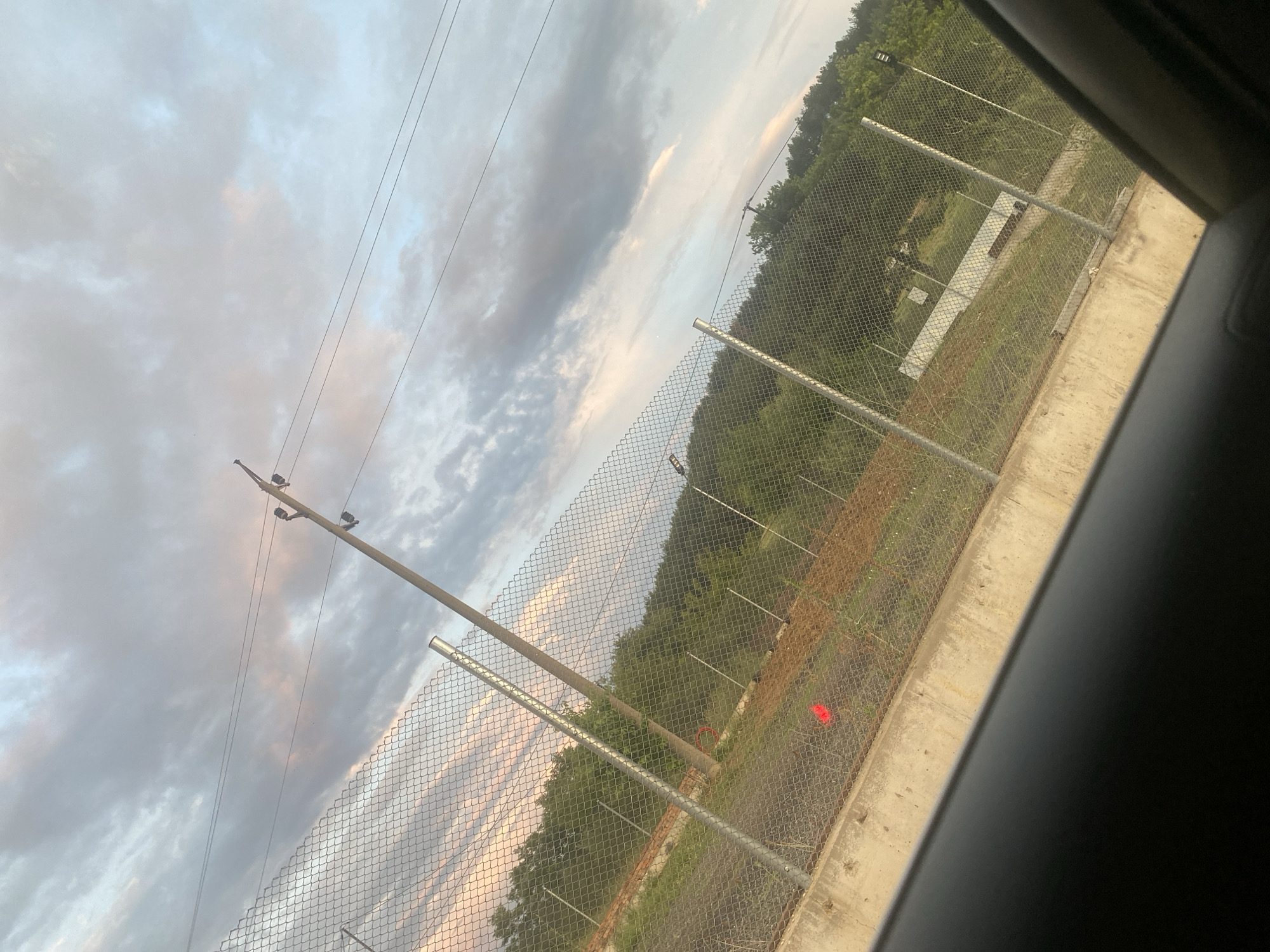 The image depicts a view through a car window of a chain-link fence, with a concrete barrier in the foreground. Beyond the fence, there are trees, a dirt area with some orange markers, and a white structure. Overhead, there are power lines and a cloudy sky with hints of pink and blue. The scene is outdoors, suggesting a roadside or perimeter setting. There are no people or animals visible in the frame. The location is specified as Ablanitsa, Bulgaria.