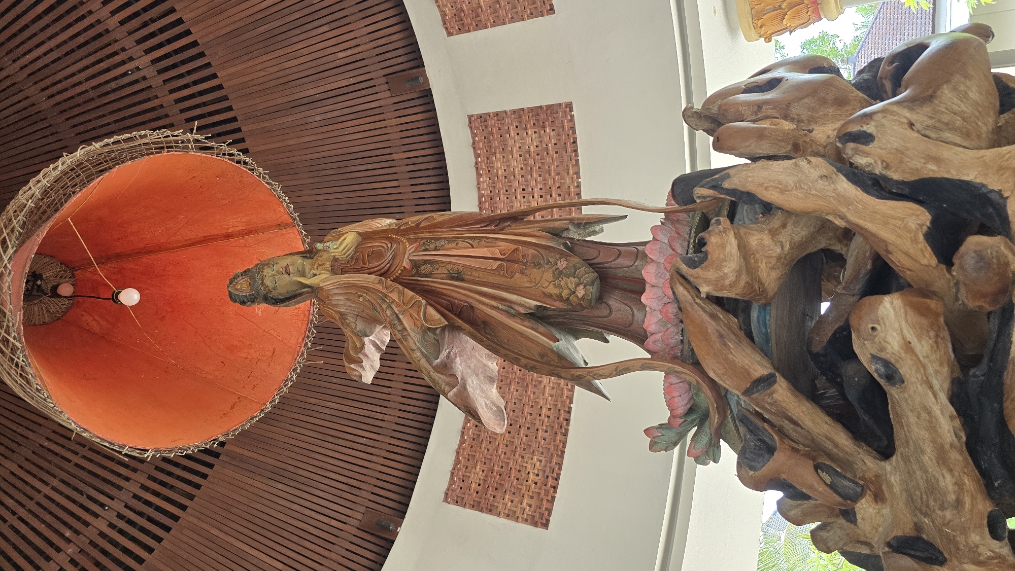 The image captures an interior scene, likely a hotel or restaurant, in the Ubud District, Indonesia. Dominating the frame are several artistic installations. A large, orange lampshade hangs from the ceiling, appearing to be crafted from woven natural materials. Attached to this lampshade is a wooden sculpture of a deity figure, possibly a Buddha or similar religious icon, carved with intricate details and floral motifs. To the right, a more abstract sculpture constructed from driftwood sits atop a white plinth. This sculpture is large and complex, featuring a variety of shapes and textures, some painted with black accents. The ceiling and walls are painted in warm brown and white tones, and have woven brick accents, creating a rustic and authentic atmosphere. The scene is brightly lit.