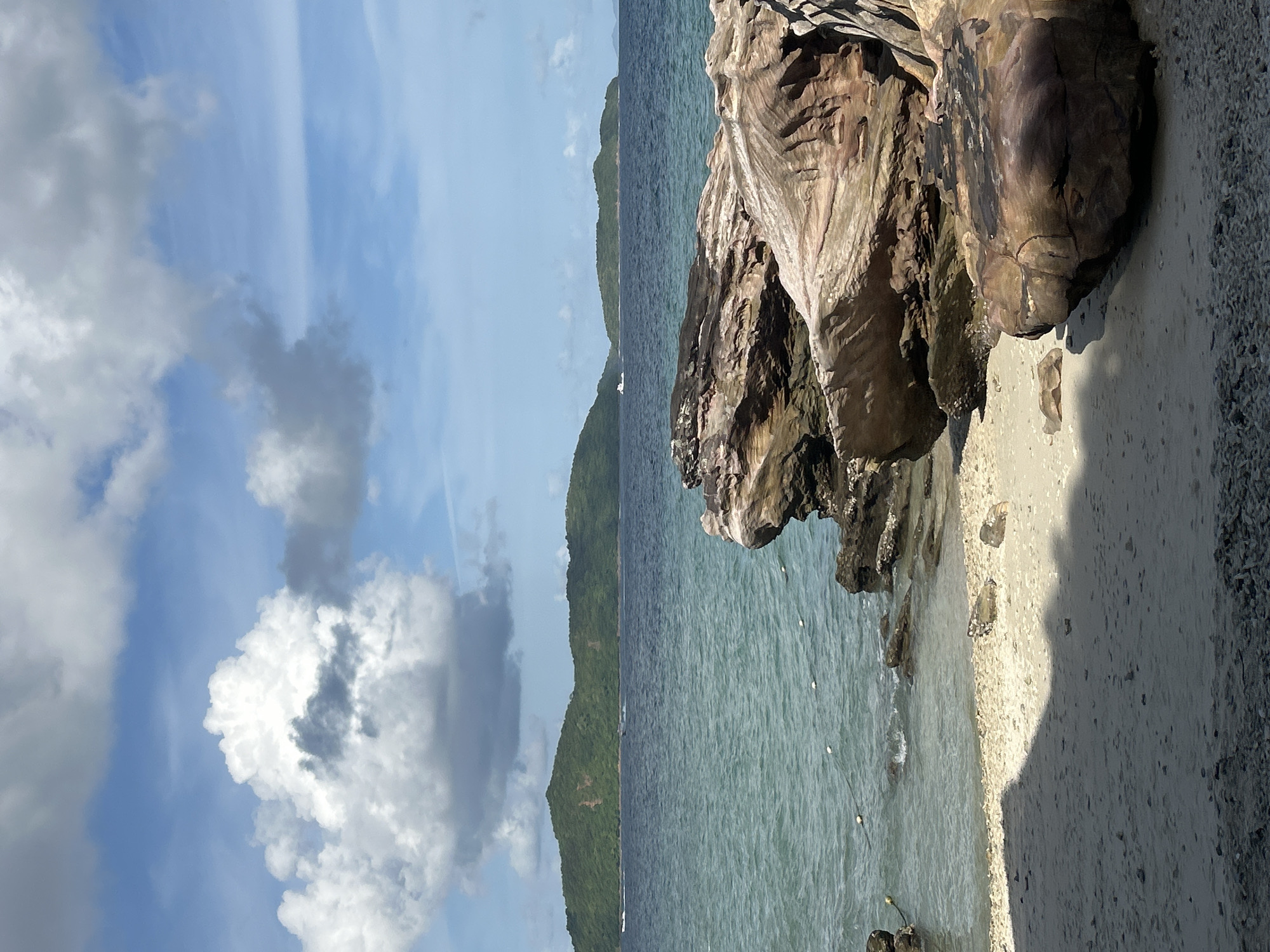 The image depicts a tropical beach scene on a sunny day, likely in Phru Nai, Thailand. The foreground is dominated by a sandy beach with some scattered pebbles and large, weathered rocks in the lower right corner. A shadow of an unseen object, possibly foliage or a structure, stretches across the sand from the left. The water is a clear, turquoise hue, gently lapping against the shore. In the middle ground, a rocky outcrop juts into the sea, with buoys visible on the water's surface. Further out, a cluster of verdant, tree-covered islands form the distant horizon. The sky is a vibrant blue, adorned with fluffy white cumulus clouds, some of which are tinged with gray. A small white boat can be seen in the distance, navigating the sea between the islands.