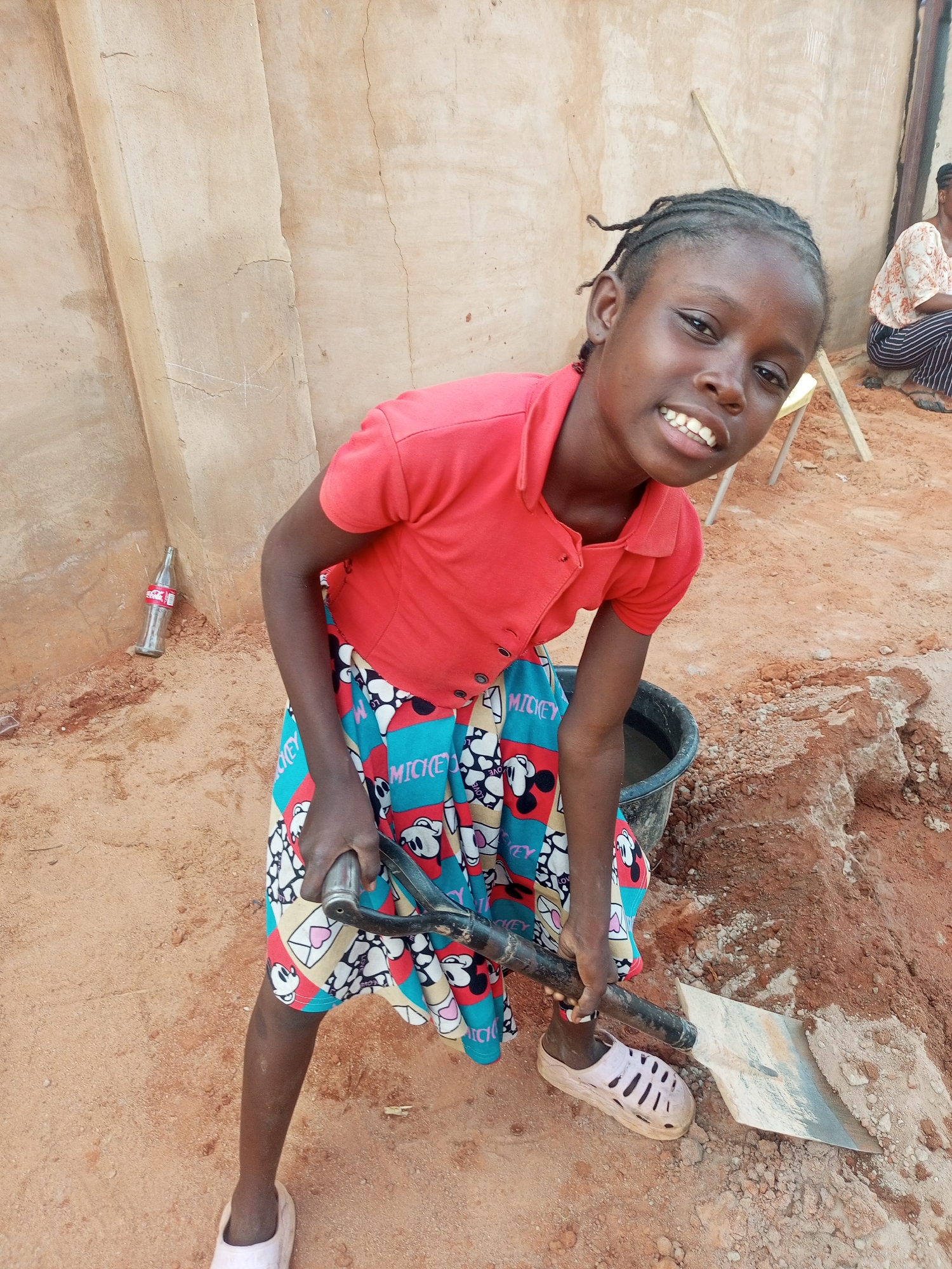 A young Black girl, likely between 8 and 12 years old, is the primary subject of this outdoor, eye-level shot taken in Tempure, Nigeria. She stands slightly hunched, facing the camera with a broad, genuine smile that reveals her teeth. Her hair is styled in dark braids.
She is dressed in a vibrant red short-sleeved collared shirt and a colorful skirt featuring a pattern of Mickey Mouse characters and hearts, with the word "MICKEY" clearly visible. On her feet, she wears light-colored, possibly plastic, sandals. In her hands, she firmly grips the black handle of a shovel or spade, the silver-grey metal blade resting on the reddish-brown soil before her. Next to her is a dark-colored bucket containing some liquid, possibly water.
The ground is predominantly reddish-brown earth, showing signs of recent excavation or disturbance, with loose soil and small rocks. The background features a light beige or sandy-colored wall, possibly made of mud or concrete, which appears somewhat weathered with visible cracks. Leaning against this wall in the mid-left is an empty glass Coca-Cola bottle, with "Coca-Cola" branding visible. Further back and to the right, a second person, an adult, is partially visible, sitting on a small chair or stool, wearing striped pants and a light-colored, patterned top. A wooden stick also leans against the wall.
The lighting suggests a clear, sunny daytime environment, with distinct shadows. The girl's cheerful expression, despite holding a tool often associated with labor, conveys a sense of happiness or playfulness.