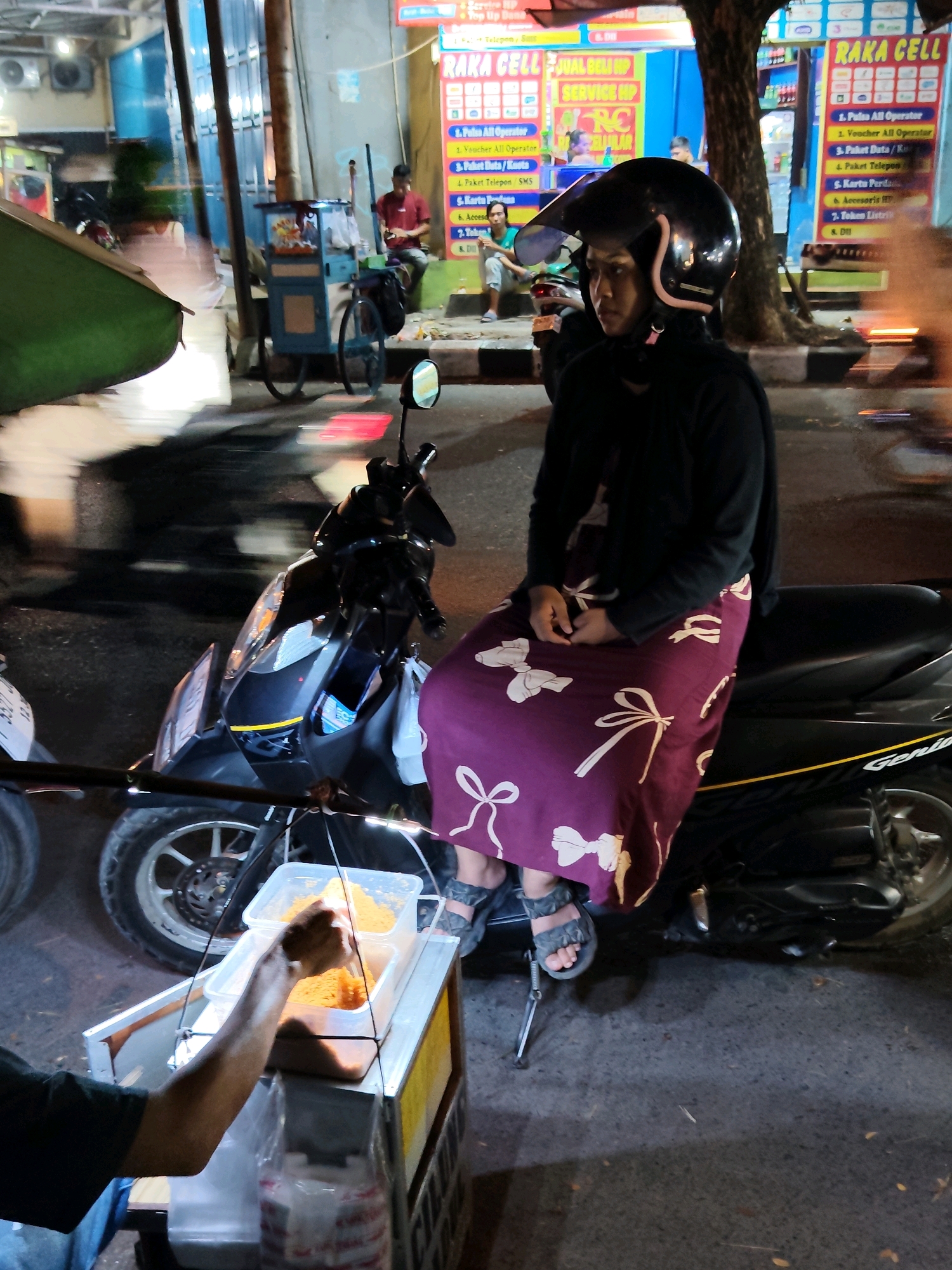 The image captures a street scene at night in Purwakarta, Indonesia, with a focus on a woman sitting on a motorcycle. She is wearing a black hijab, a black jacket, and a long purple skirt adorned with white bow patterns. Her feet are clad in grey sandals. The motorcycle she is seated on is black and parked on a street with a paved surface.
In the foreground, a food cart is visible, with a hand reaching into a transparent container filled with orange-colored food items, likely snacks or ingredients. The cart has a metal frame and a yellow trim.
Behind the woman and motorcycle, the background reveals a shop with illuminated signs, one of which reads "RAKA CELL" in large letters, suggesting it's a mobile phone or telecommunications store. The shop's facade is brightly lit, contrasting with the darker street. Other elements in the background include trees, streetlights, and blurred figures of people. The overall atmosphere suggests a bustling street at night, possibly near a commercial area.