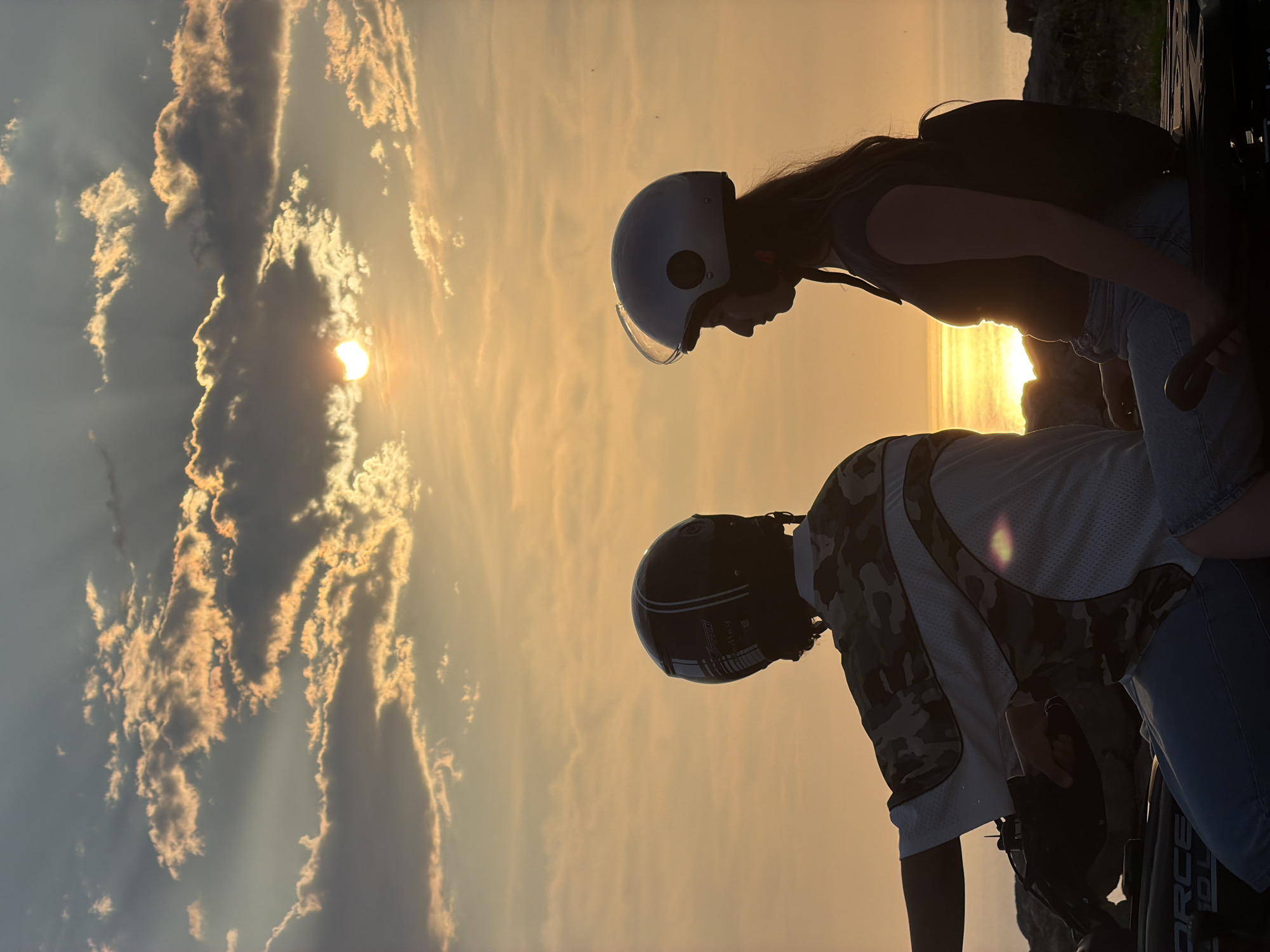 In Kovačko Polje, Montenegro, two individuals, a male and a female, are seated on an ATV, silhouetted against a dramatic sunset. The sky is filled with a mix of dark and golden clouds, with rays of sunlight breaking through, illuminating the scene with a warm, orange glow. The sun itself is partially obscured by clouds, positioned in the upper center of the frame. The male, in the foreground, wears a camouflage patterned shirt and a black helmet. The female, seated behind him, is wearing a dark top and a silver helmet. Both are looking out towards the horizon. The ATV is dark-colored with some visible lettering. The background suggests a rocky, natural landscape meeting the water, which reflects the sunset.