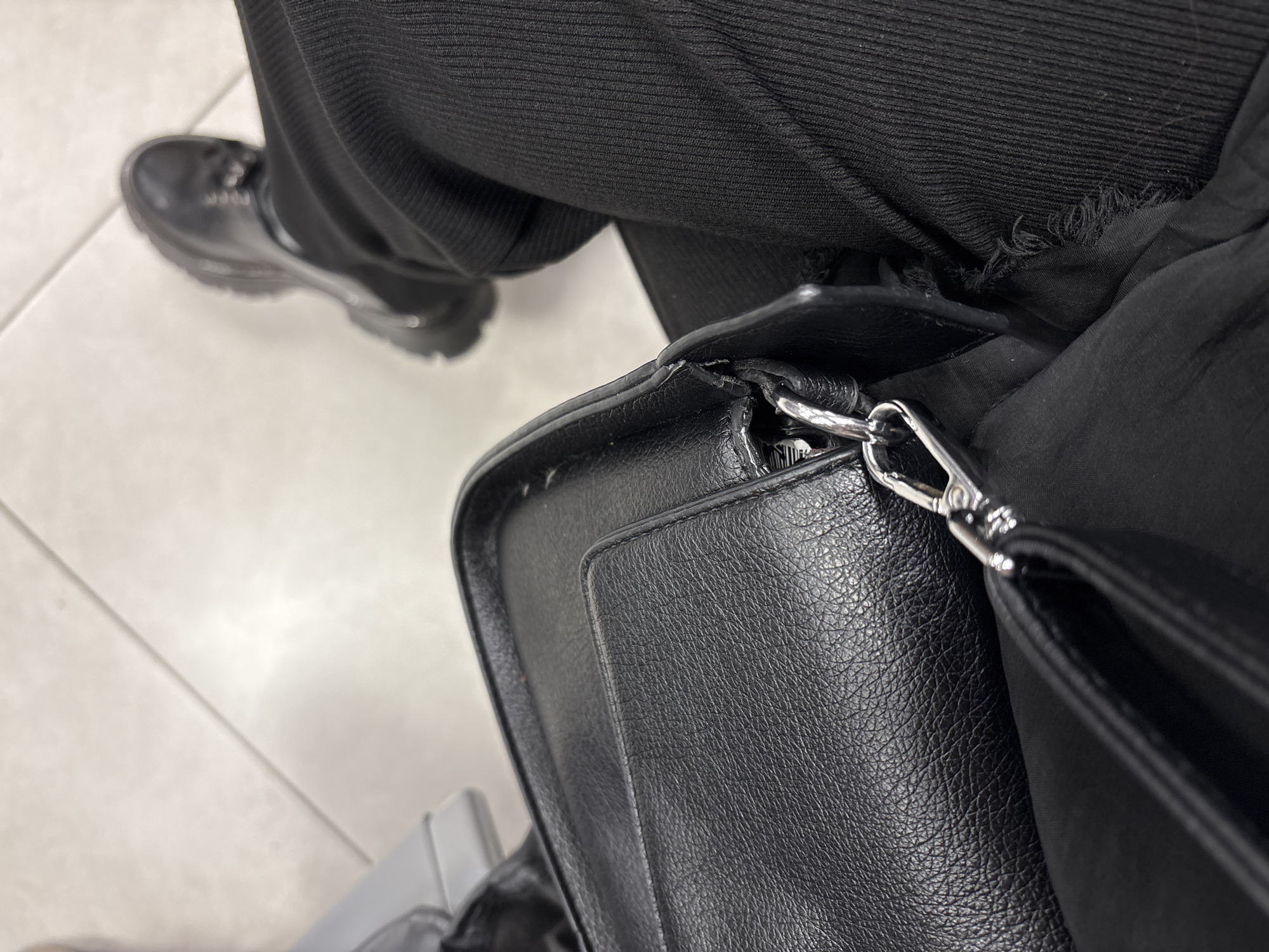 A high-angle, top-down shot captures the lower portion of a person's body and a bag, set against an indoor tiled floor. The foreground is dominated by a black, textured bag, appearing to be faux leather, with a prominent silver-toned metal swivel snap hook. A seam and what appears to be an outer pocket or flap are visible on the bag's surface. To the right, a person's right leg is clad in black trousers, featuring a visible ribbed or corduroy-like texture and a distressed or torn hem near the bag. Further back, the person's right foot is encased in a black, chunky-soled boot with visible laces and a thick, ridged sole. The floor is composed of light-colored, possibly off-white or light grey tiles, with distinct grout lines forming a geometric pattern. A bright reflection or light source illuminates a section of the floor in the upper right. A blurry, light-toned object is partially visible in the upper left corner. This scene is located in the city of Bol'shie Kabany, Russia.