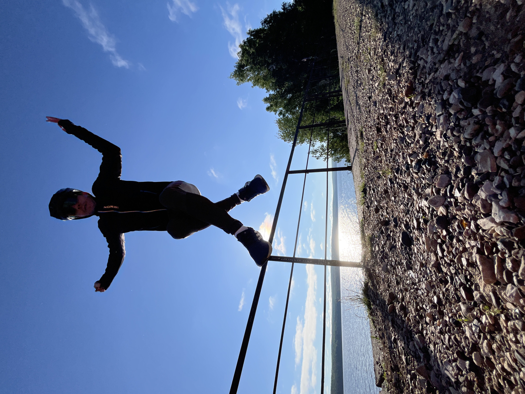 A low-angle shot captures a person in mid-air, jumping with legs crossed and arms outstretched, against a bright blue sky with scattered clouds. The individual is wearing dark clothing, a helmet, and sunglasses. They appear to be leaping over a black metal railing that runs horizontally across the frame. Below the railing, a body of water, likely a lake or river, reflects the sun. The foreground is covered in pebbles and rocks, with some sparse green vegetation. Trees are visible in the background on the right side of the image. The scene suggests an outdoor recreational area, possibly near the Kama River in Chaikovsky, Russia, given the presence of water and the natural landscape.