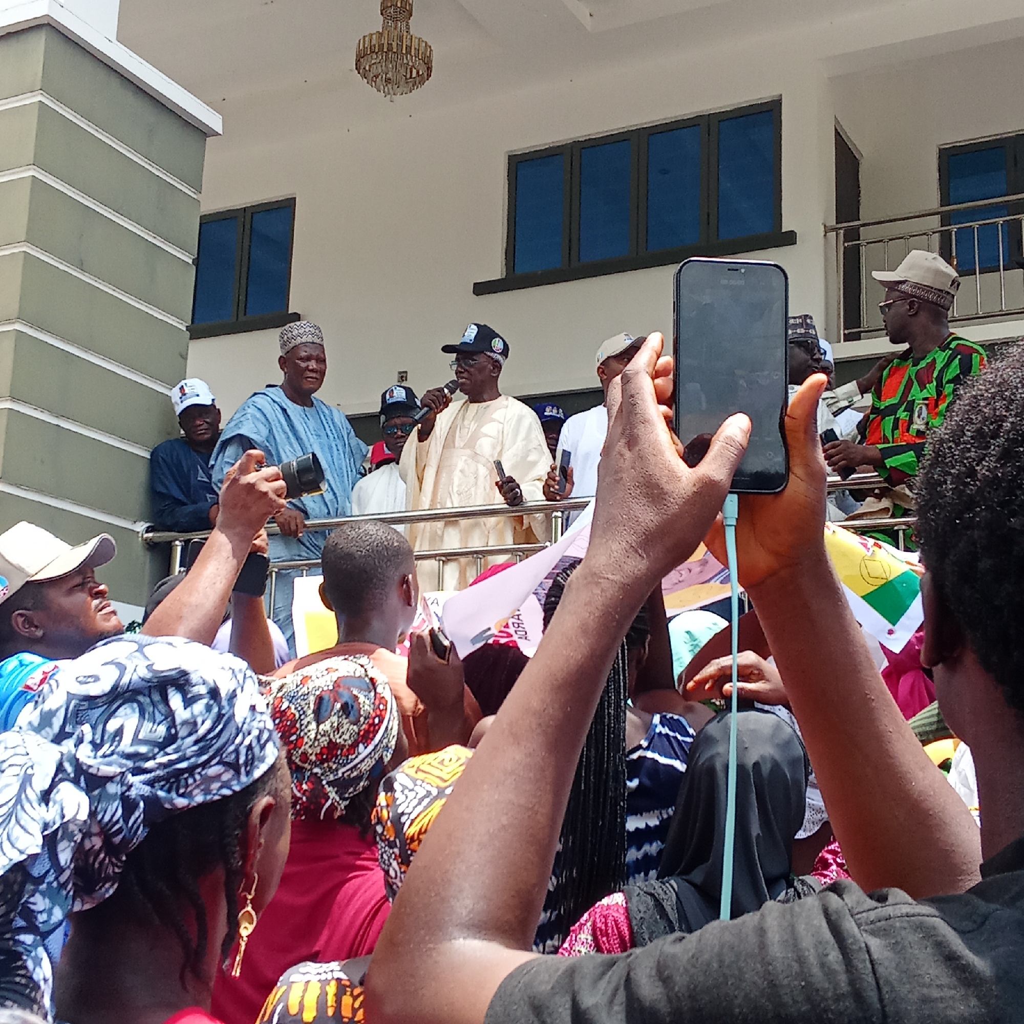 A crowd gathers outside a white building with a green-striped column in Akwanga, Nigeria. Several men stand on a raised balcony speaking into microphones. The crowd below holds flags and cell phones, capturing the event. The sky is bright and the building features a chandelier hanging near the balcony.