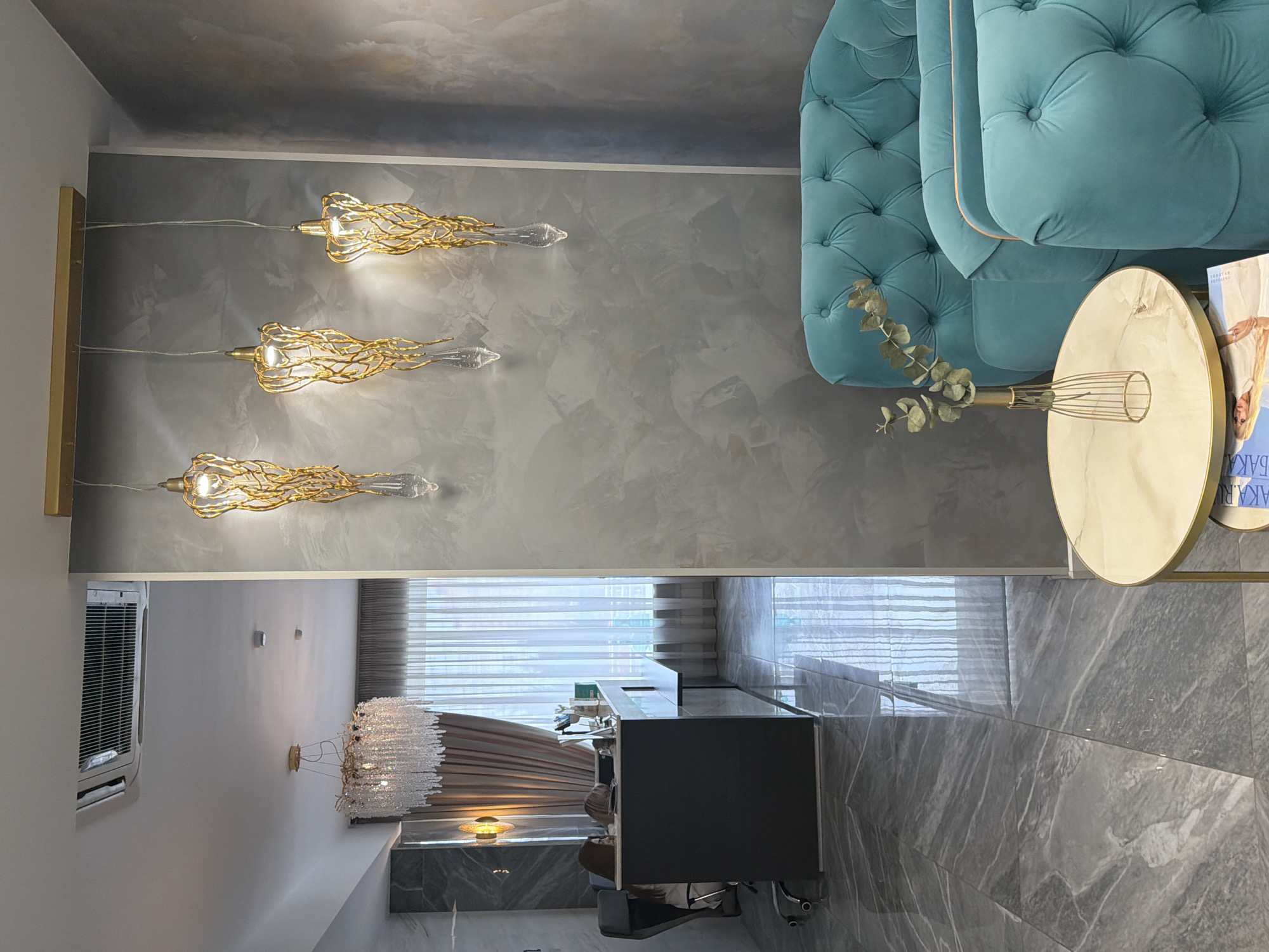 A vertically-oriented interior shot captures a modern space, likely within an apartment or house. The color palette is dominated by shades of gray, gold, and a muted teal. The photograph is taken from a relatively high angle, looking down on the scene.
On the right, a portion of a teal tufted sofa is visible, featuring an accent side table with a circular, light-colored top and a gold frame. Adjacent to this, a vase holds sprigs of eucalyptus.
The wall on the left is painted a textured gray, with three gold-toned pendant lights arranged vertically. These lights have intricate, flowing designs with clear glass tips.
Extending into the background, a modern bathroom or kitchen area is visible. A dark gray countertop or island sits on a marble-tiled floor. Further back, a window covered with sheer white blinds is illuminated by warm light. A chandelier-style light fixture hangs above, consisting of numerous small, clear crystals.
While it's difficult to confirm the exact location without additional information or context, the image's style and furnishings suggest a modern residential space, potentially located in a city like Krasnoyarsk, Russia. The architecture and interior design elements are consistent with contemporary trends. However, more precise geolocation data would be necessary for confirmation.
