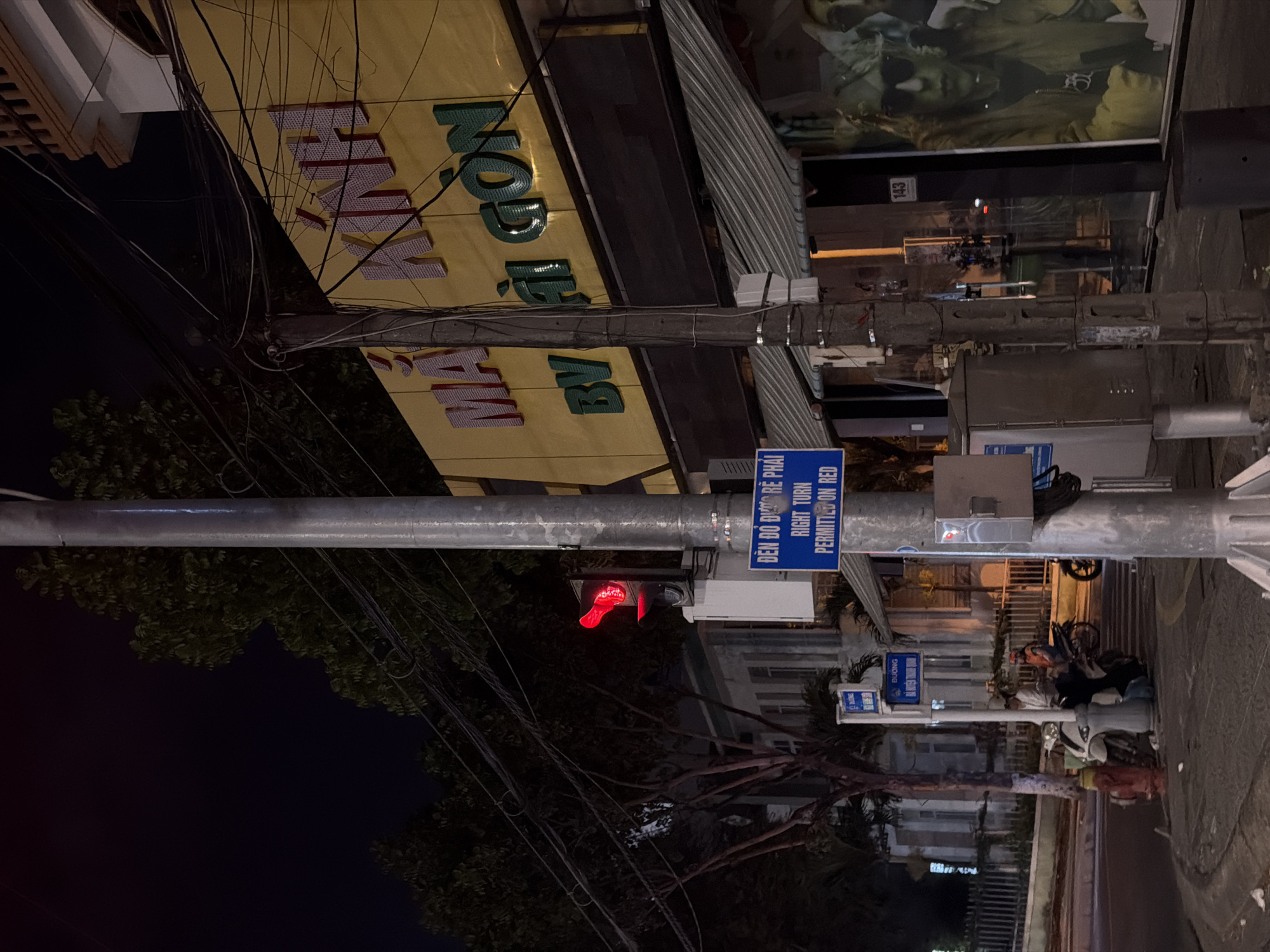 The image captures a street scene at night in Đà Nẵng, Vietnam. A traffic light is visible, displaying a red signal, with a sign underneath stating "DEN DO DUOC RE PHAI RIGHT TURN PERMITTED ON RED". A street pole stands prominently in the foreground, supporting the traffic light and numerous electrical wires. To the right, a building with a yellow facade features large, raised letters spelling out "MAI KINH" and "BY AI GON". A storefront window displays a poster of a person wearing sunglasses. In the background, trees and a building with a fence are visible. The street appears wet, reflecting the ambient light. Several motorcycles and people are present, indicating ongoing activity.