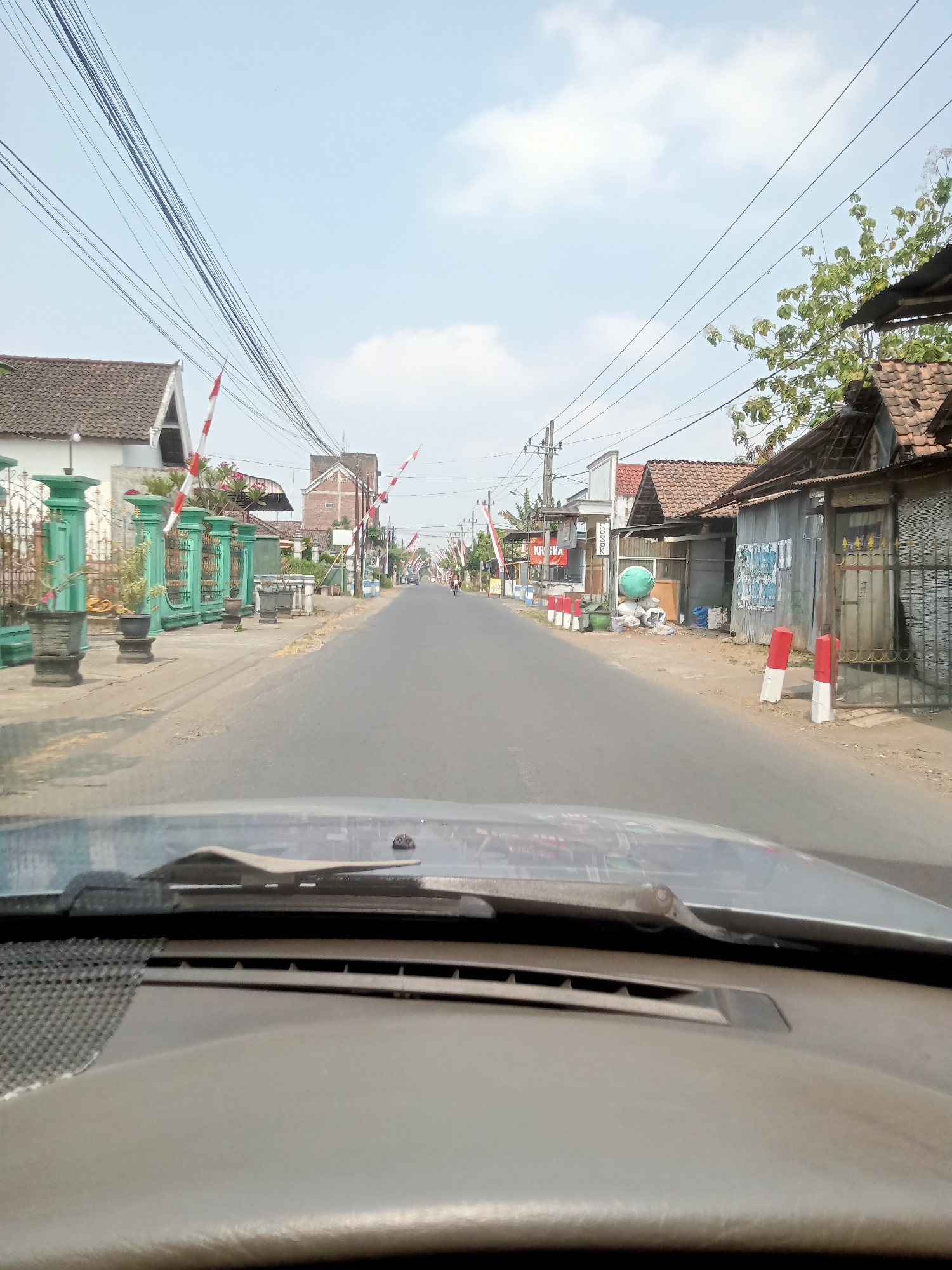 A first-person perspective from a vehicle looking forward along a paved road in Majan, Indonesia. The road runs through a residential area with buildings lining both sides. Houses are visible, some with tiled roofs and others with corrugated metal. Red and white striped barriers are positioned along the roadside, suggesting a crossing, possibly a railroad. Power lines are strung overhead, supported by utility poles. The sky is blue with scattered clouds. On the left side of the road, there is a green fence with decorative pillars and potted plants. On the right, there are more structures, including some appearing to be shops or small businesses, with various items piled outside, such as sacks and a large green cylindrical object. Further down the road, a person is seen riding a motorcycle. The scene appears to be set during daylight.