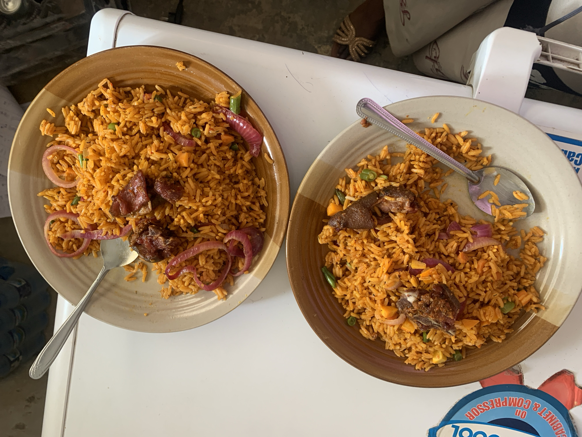 Two plates of jollof rice are on a white surface, likely a table or countertop. The rice dishes appear to be topped with pieces of meat and red onion slices. Each plate has a spoon in it. The image appears to be taken indoors, possibly in a home or restaurant setting. The background includes parts of furniture or appliances. The location is unknown.