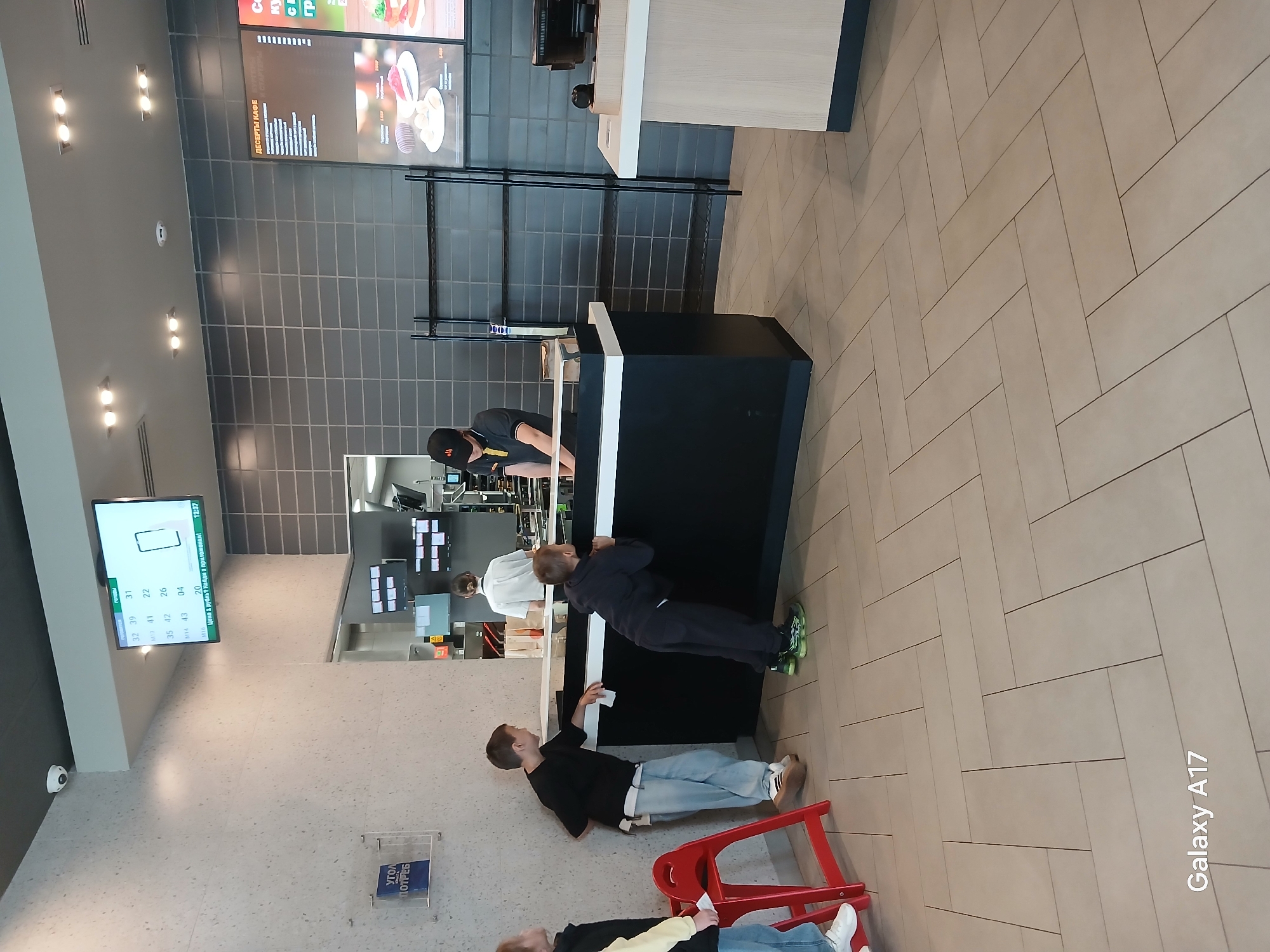 Here is a matter-of-fact description of the image:
The image shows the interior of a fast-food restaurant, likely a McDonald's, in the city of Yekaterinburg, Russia. The interior features tiled floors, a counter area with a black and white design, menu boards displaying food options and prices, and digital displays above the counter showing order information. Several people are visible, including employees behind the counter and customers waiting to order or receive their food. Some customers are standing near the counter, while others are seated on red plastic chairs. A sign with Cyrillic text is mounted on the wall. The image seems to have been captured by a phone, based on the 'Galaxy A17' watermark in the bottom right corner.