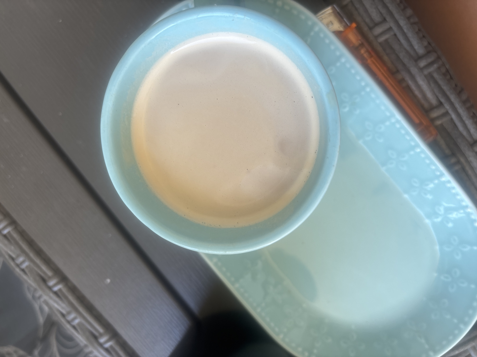 A close-up, overhead shot captures a light blue mug filled with coffee, its surface showing a thin layer of foam, resting on a matching light blue plate with a subtle embossed floral pattern. The plate is positioned on a grey, woven outdoor furniture surface. An orange object, possibly a lighter or pen, lies on the plate near the mug. The scene is set outdoors, with the wicker furniture suggesting a patio or balcony setting. There is no indication of the location being in Plovdiv, Bulgaria.