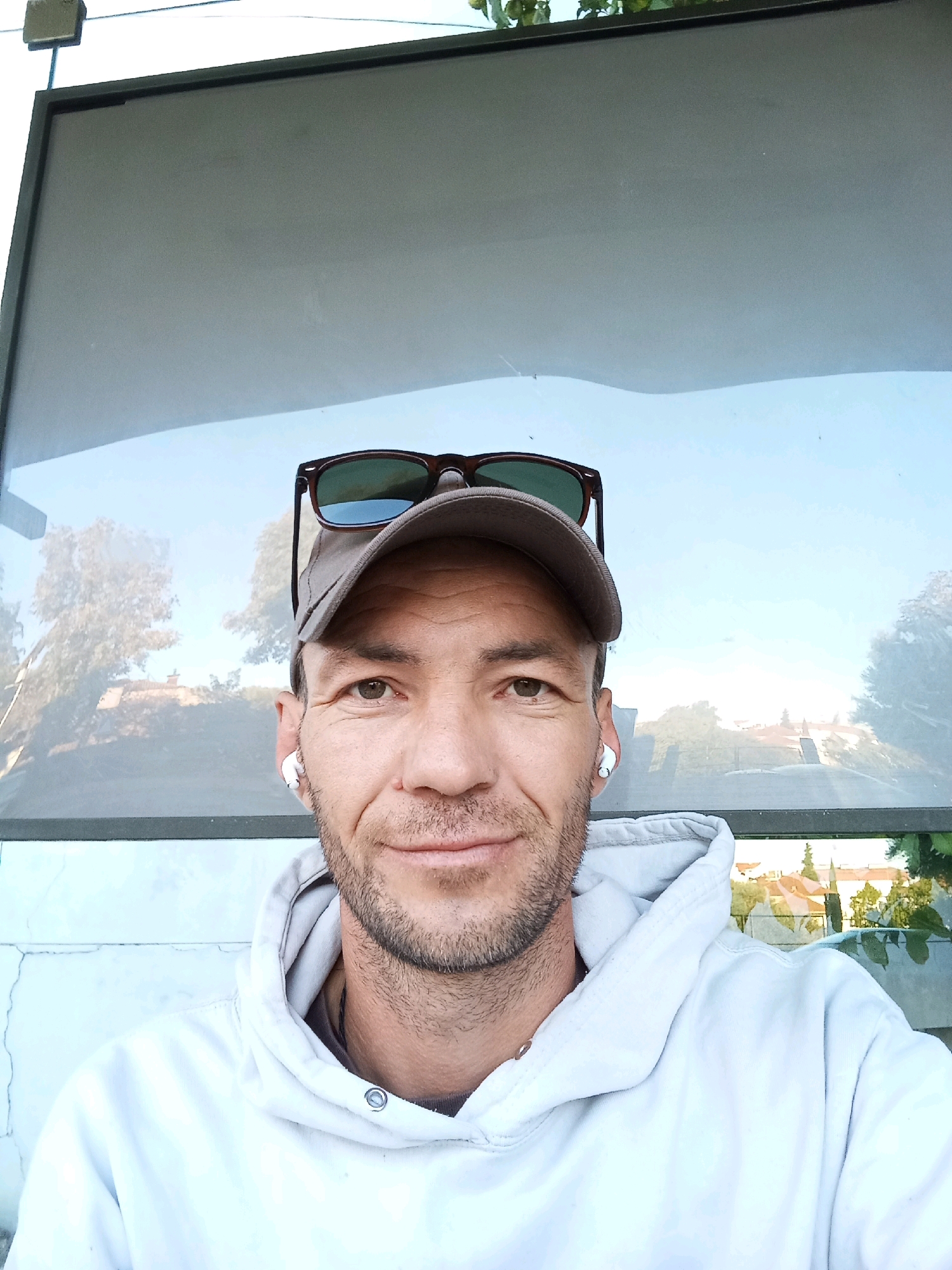 A man with a baseball cap and sunglasses is taking a selfie outdoors. He is wearing a light-colored hoodie and has white earbuds in his ears. The background is a reflective surface showing trees and buildings, suggesting a public area or possibly a bus stop. The man has a short beard and is looking directly at the camera with a slight smile. The lighting suggests it is daytime. It is not possible to determine the exact location in Viseu, Portugal, from the image alone.
