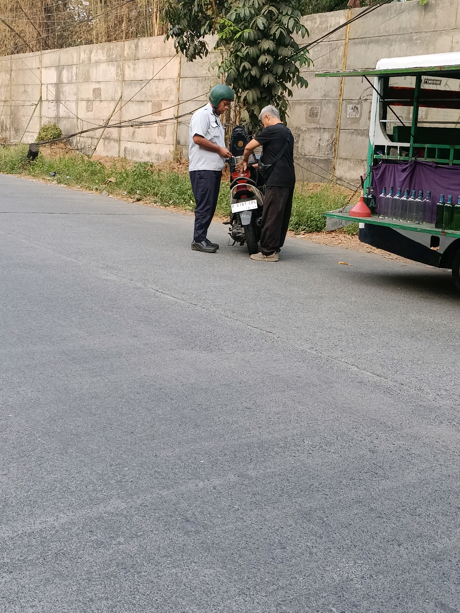 Two men are standing on a paved road next to a motorcycle and a small truck. The man on the left is wearing a light blue short-sleeved shirt, dark pants, and a green helmet. He appears to be holding a white object. The man on the right is wearing a black t-shirt and dark pants. He is bending over and appears to be interacting with the motorcycle. The motorcycle is red and black and has a license plate that reads "B 6787 CPG".
The truck is green and white and has a canopy. Several glass bottles, some empty and some filled with a clear liquid, are arranged on the back of the truck. A red funnel is also visible on the truck bed. The truck has a sign on its side with Indonesian writing.
The scene is set against a concrete wall with some vegetation in front of it. Overhead wires are visible above the wall. The overall environment suggests an outdoor setting, possibly in a semi-urban or rural area of Tangerang, Indonesia, given the visible license plate format and the type of vehicle. The lighting suggests it is daytime.