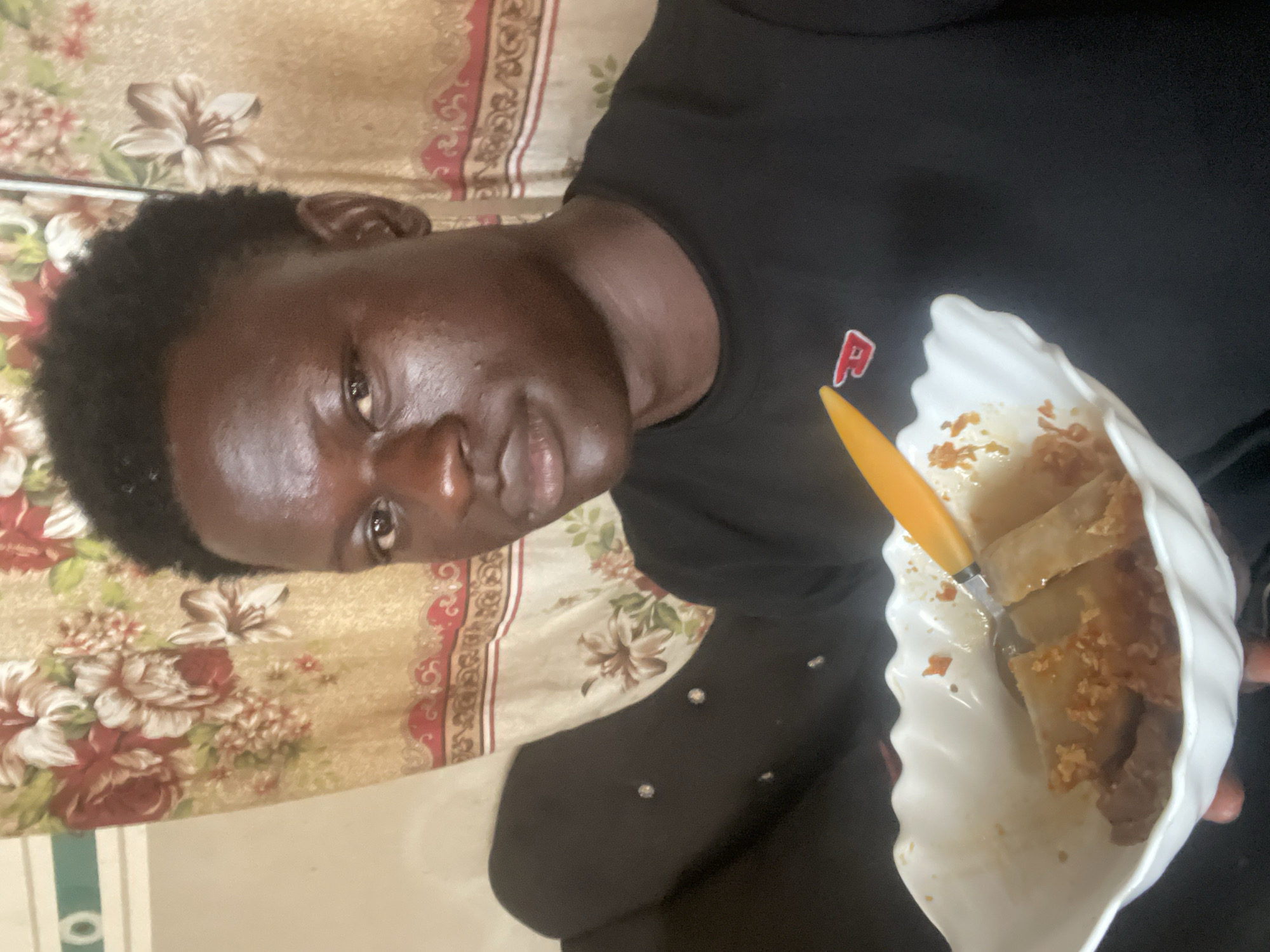 A person with dark skin and short dark hair is holding a white, scalloped dish with food in it. The person is wearing a black shirt with a red letter "P" on it. A yellow plastic utensil is in the dish. The background includes a floral patterned curtain or blanket. The person is in a room with a beige wall. The location appears to be Badagry, Nigeria.