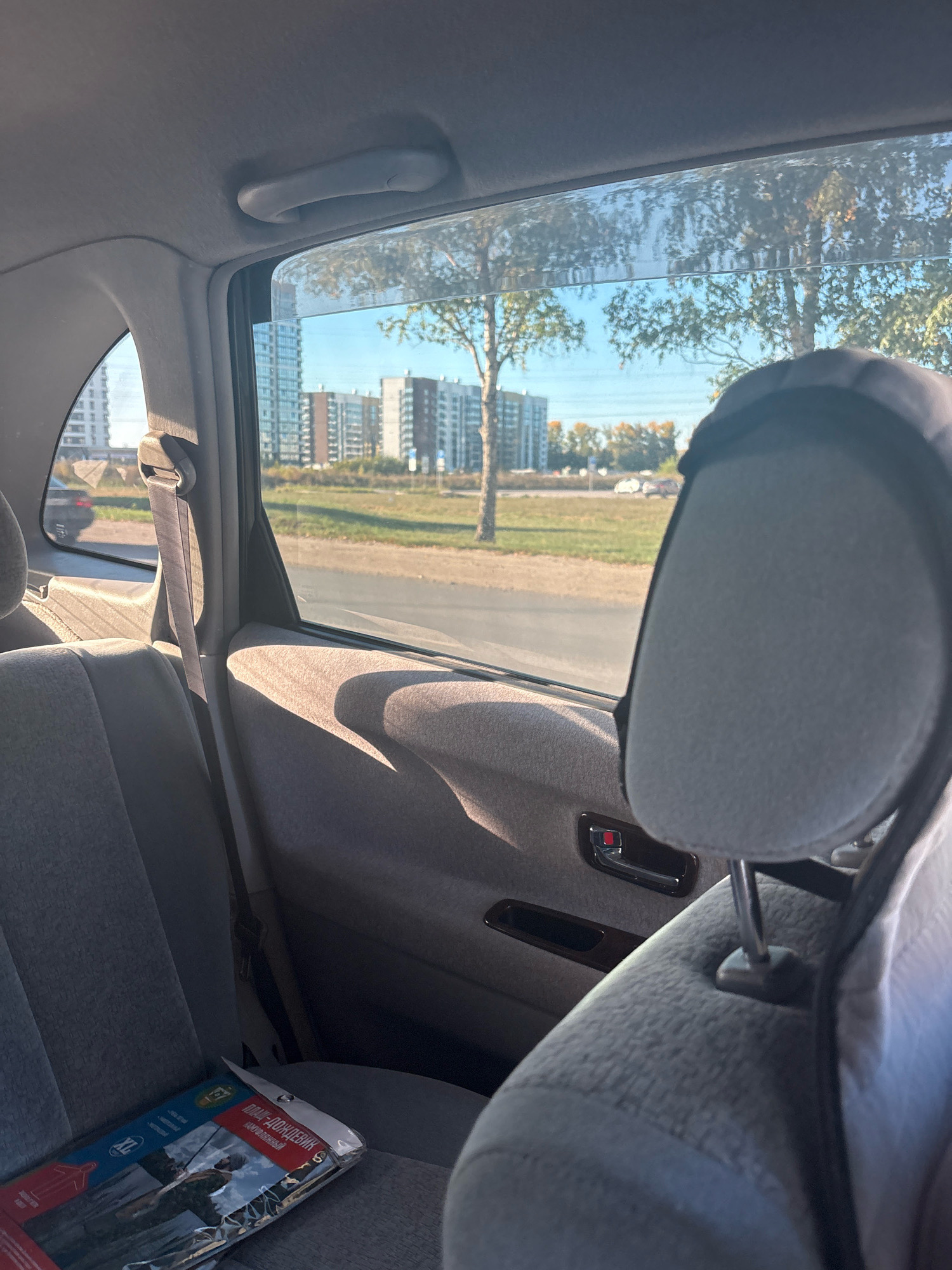 This is a photograph taken from inside a vehicle, looking out the passenger-side window. The interior of the car is visible in the foreground, including gray cloth seats, a headrest, and a seatbelt. On the seat is a package with Russian text, the title visible appears to be "ДОЖДЕВИК" which translates to "raincoat". Outside the window, a tree is visible, along with several multi-story buildings in the distance. The sky is blue with some clouds, and sunlight is casting shadows into the car. The location is the city of Barnaul, Russia.