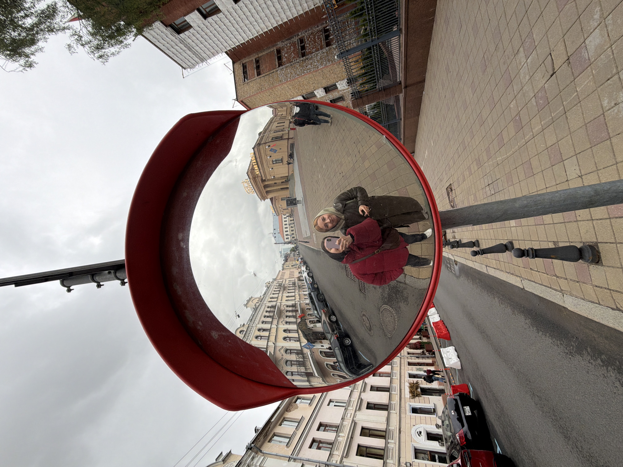 A convex traffic mirror mounted on a pole reflects a street scene. Two individuals are captured in the mirror's reflection, positioned closely together and facing the camera, indicating they are likely taking a selfie. One person is wearing a red coat, while the other is wearing a dark coat with a hood. The reflection also shows an urban street with parked cars, buildings with traditional architecture, and a paved pedestrian area. The sky is overcast. The location appears to be in Unknown, Unknown.