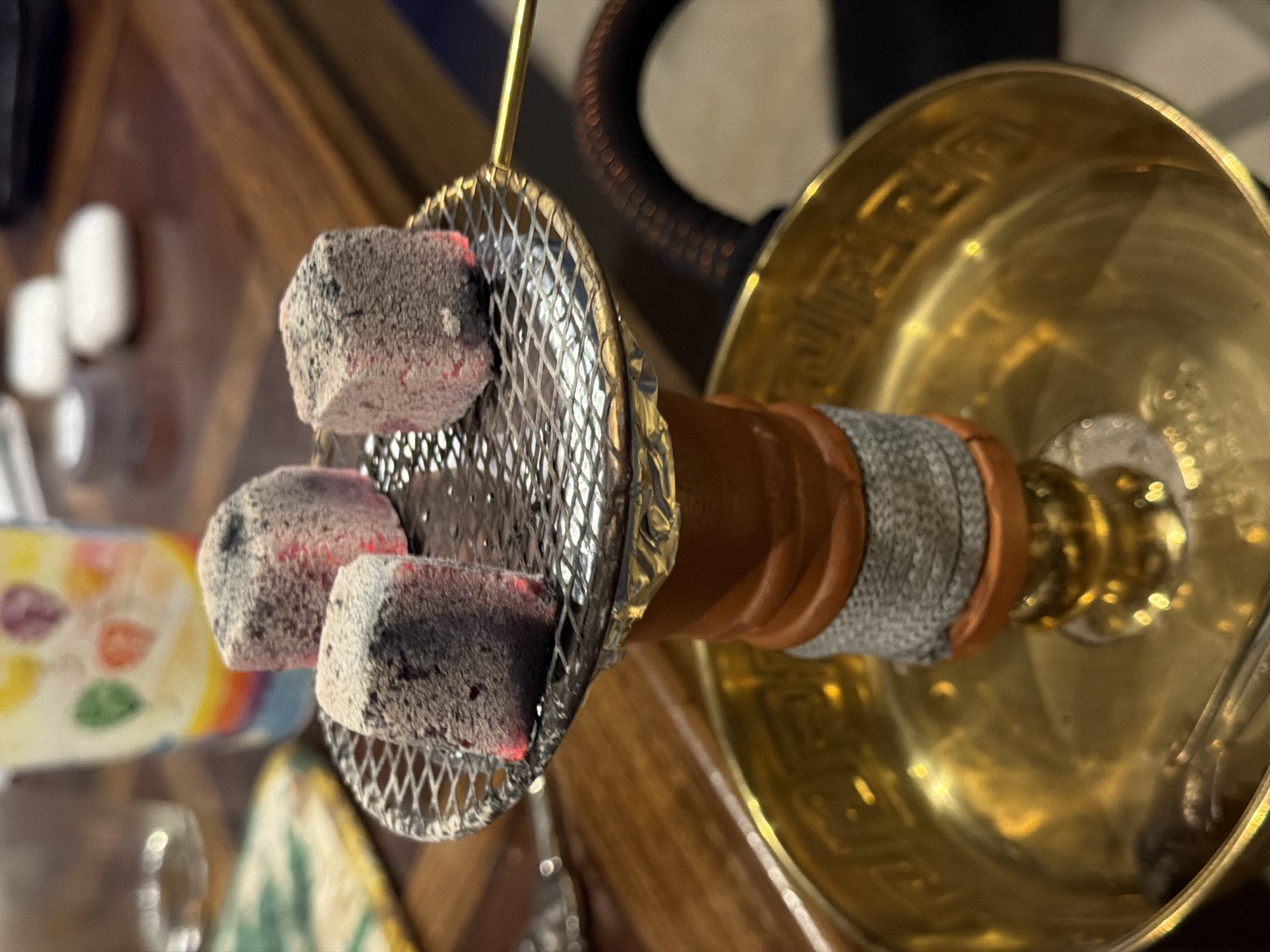 In Sarouja Municipality, Syria, a close-up shot focuses on a hookah setup. Three charcoal briquettes, glowing red from within and dusted with ash, rest on a circular metal screen. This screen sits atop the hookah's heat management system, which is constructed from orange-brown ceramic and wrapped in gray decorative fabric. Below this, the wide, ornate brass bowl of the hookah gleams, reflecting the ambient light. The background is blurred but suggests a wooden table surface with other objects, including a colorful, patterned cup or vase and a dark, possibly electronic device. The lighting is dim, highlighting the red glow of the coals.
