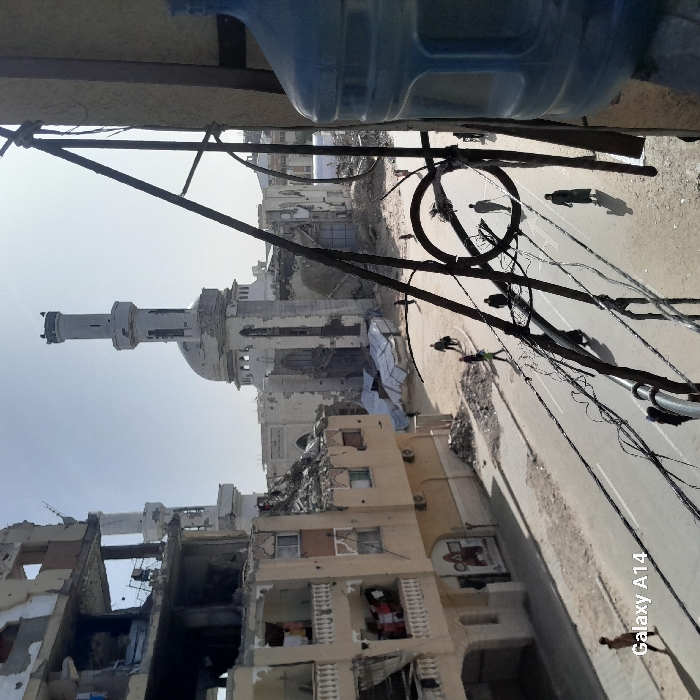The image depicts a scene of urban destruction, likely in the Middle East, possibly Gaza. Buildings are heavily damaged with visible structural collapse and rubble. A mosque with a prominent minaret stands amidst the devastation. Utility poles and wires clutter the view. The streets appear desolate, with a few figures visible in the distance. The photo's tilt gives a disorienting perspective. A blue water jug sits atop the frame. Overall, the image conveys the aftermath of conflict and the profound impact on civilian infrastructure.