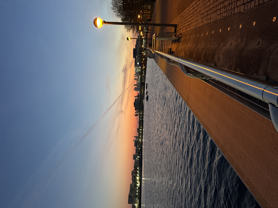 The image depicts an outdoor waterfront scene at dusk, likely in the Royal Victoria Dock area of the Royal Docks in East London, United Kingdom.
The sky transitions from deep blue at the top to a vibrant orange and red at the horizon, indicating sunset or twilight. Two prominent, faint contrails from aircraft are visible diagonally across the upper sky. A wide body of water, likely the Royal Victoria Dock, occupies the left half of the frame, its surface dark blue-grey with subtle ripples, reflecting the orange glow of the sky and artificial lights.
In the foreground, running along the right side of the image, is a waterfront promenade. Immediately adjacent to the water is a textured, orange-brown walkway surface. A low, shiny metallic railing extends horizontally along this edge, reflecting the warm artificial light. Beyond the railing, a paved walkway of cobblestone-like material is visible, illuminated by several circular ground lights embedded into its surface. A tall, dark streetlight with a spherical, warm yellow-orange glowing fixture stands prominently on the far right, with other similar streetlights receding into the distance along the promenade.
Across the water, on the far bank, a continuous line of illuminated buildings, ranging from residential to commercial structures, forms the mid-ground skyline. Their windows and exterior lights are active. Several construction cranes are discernible among these buildings. On the water, in the mid-ground towards the left, at least three small, dark boats, possibly recreational rowboats or pedal boats, are present.
Further along the promenade in the mid-ground right, indistinct figures of several people can be seen walking, silhouetted against the ambient light from the streetlights and ground lights. Sparse, bare tree branches are visible on the extreme right, suggesting deciduous trees without foliage. The distant skyline further east or west of the Royal Docks is visible as a collection of faint lights and building silhouettes against the colorful horizon.