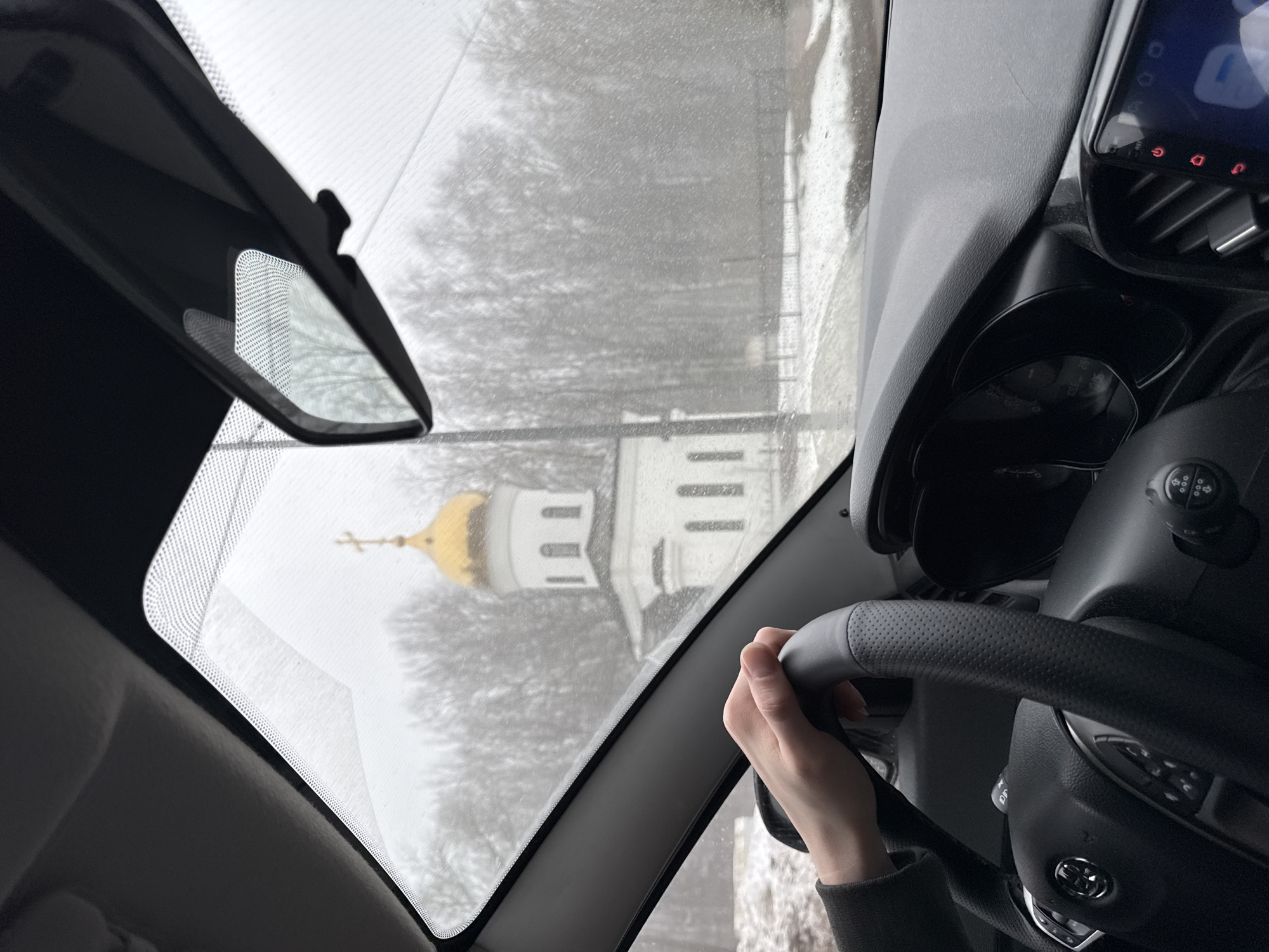 The photograph is taken from inside a car, looking out through the windshield, capturing a view of a church in Votkinsk, Russia. The church features a golden dome and white walls, situated amidst leafless trees, suggesting a cold season. The windshield is slightly obscured by what appears to be rain or snow. Inside the car, a hand is visible on the steering wheel, indicating a person driving or about to drive. The dashboard and rearview mirror are also visible, providing context to the car's interior. The overall environment seems overcast and muted due to the weather conditions.