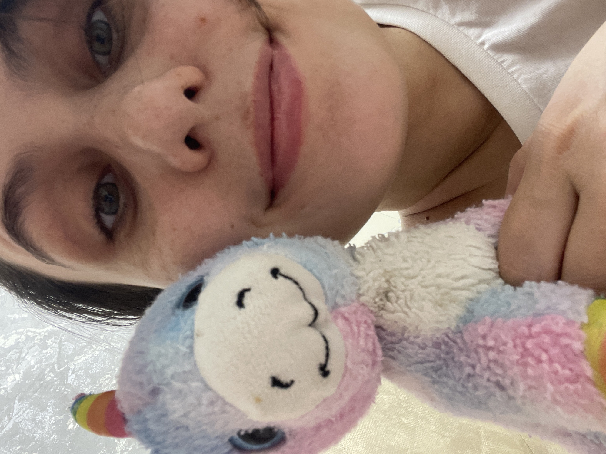 The image presents a close-up, high-angle view of an individual holding a plush toy in an indoor setting. A female's face occupies the right half of the frame, showing dark hair, medium-dark eyes, a nose, and closed lips with a subtle upward curve. Her skin displays freckles. She is wearing a white t-shirt with a visible ribbed collar on the lower right. In the foreground, partially obscuring the person's left side, is a multi-colored plush toy, appearing to be a unicorn or pony due to a rainbow-striped horn on its head. The toy features pastel shades of pink, blue, purple, and white fur, with black bead eyes and a stitched black smiling mouth on a white snout. The individual's hand supports the toy. The background is a uniformly light, textured surface, providing no specific environmental details. The exact location within Izhevsk, Russia, cannot be determined from the visual evidence in the image.