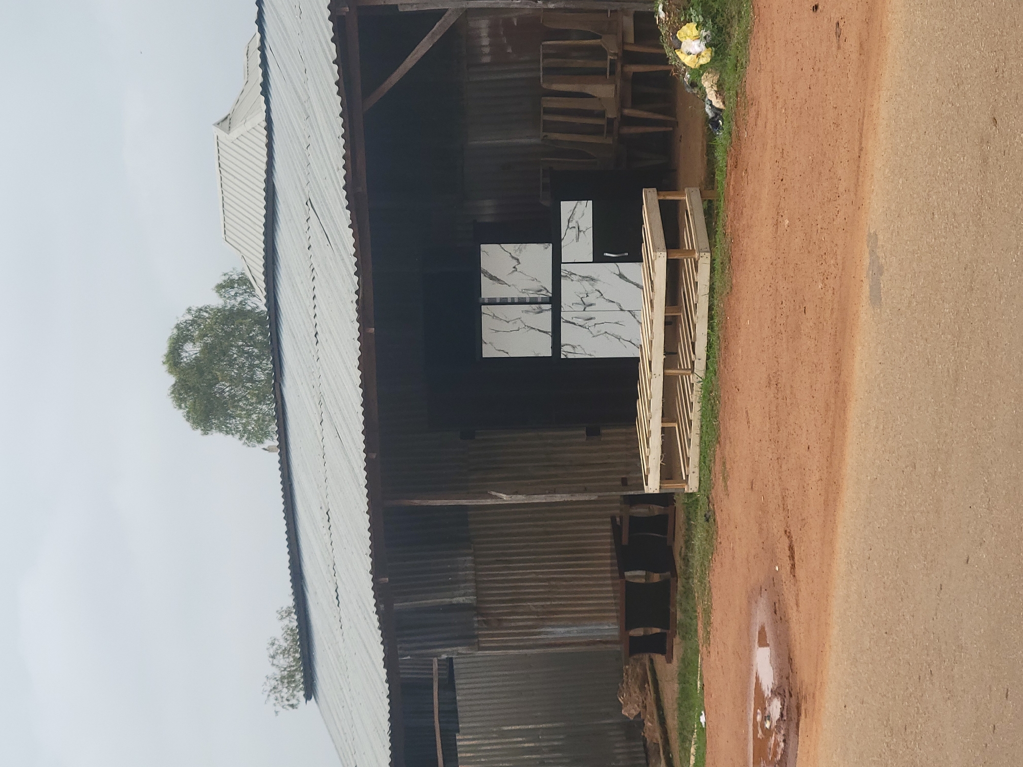 The image depicts an outdoor scene in front of a corrugated metal structure, possibly a shop or workshop, in Barakin Akawo, Nigeria. The structure has a weathered corrugated metal roof supported by wooden beams. In front of the structure, on a dusty, reddish-brown ground, are several items for sale or display. These include a wooden pallet rack, two dark-colored bench-like objects, and a dark cabinet unit with white marble-patterned panels and a silver handle. Behind the cabinet, several wooden chairs are stacked. To the right of the structure, there is a pile of trash, including yellow and white plastic bags. A muddy puddle is visible on the ground to the left of the structure. The sky is overcast and light gray. The overall impression is of a humble commercial establishment in a semi-rural or unpaved urban setting. There are no people visible in the image.