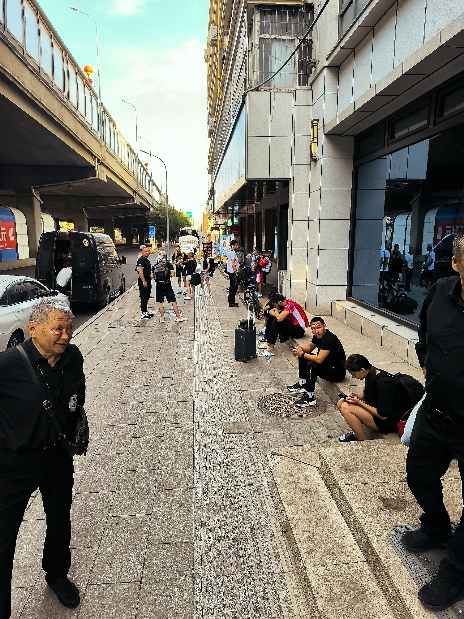 An elderly Asian man in a black shirt and pants walks past a sidewalk in Ürümqi, China, smiling towards the left side of the frame. He carries a black crossbody bag. To his right, on the sidewalk, a group of people are gathered. Several individuals are sitting on the curb in front of a building with a reflective glass facade, looking at their phones. One person is sitting with their head in their hands. A black rolling suitcase is on the ground near them. In the background, a black van is parked with its rear doors open, and a white car is parked in front of it. Further down the street, an elevated highway overpass is visible. The sky is bright with some clouds.