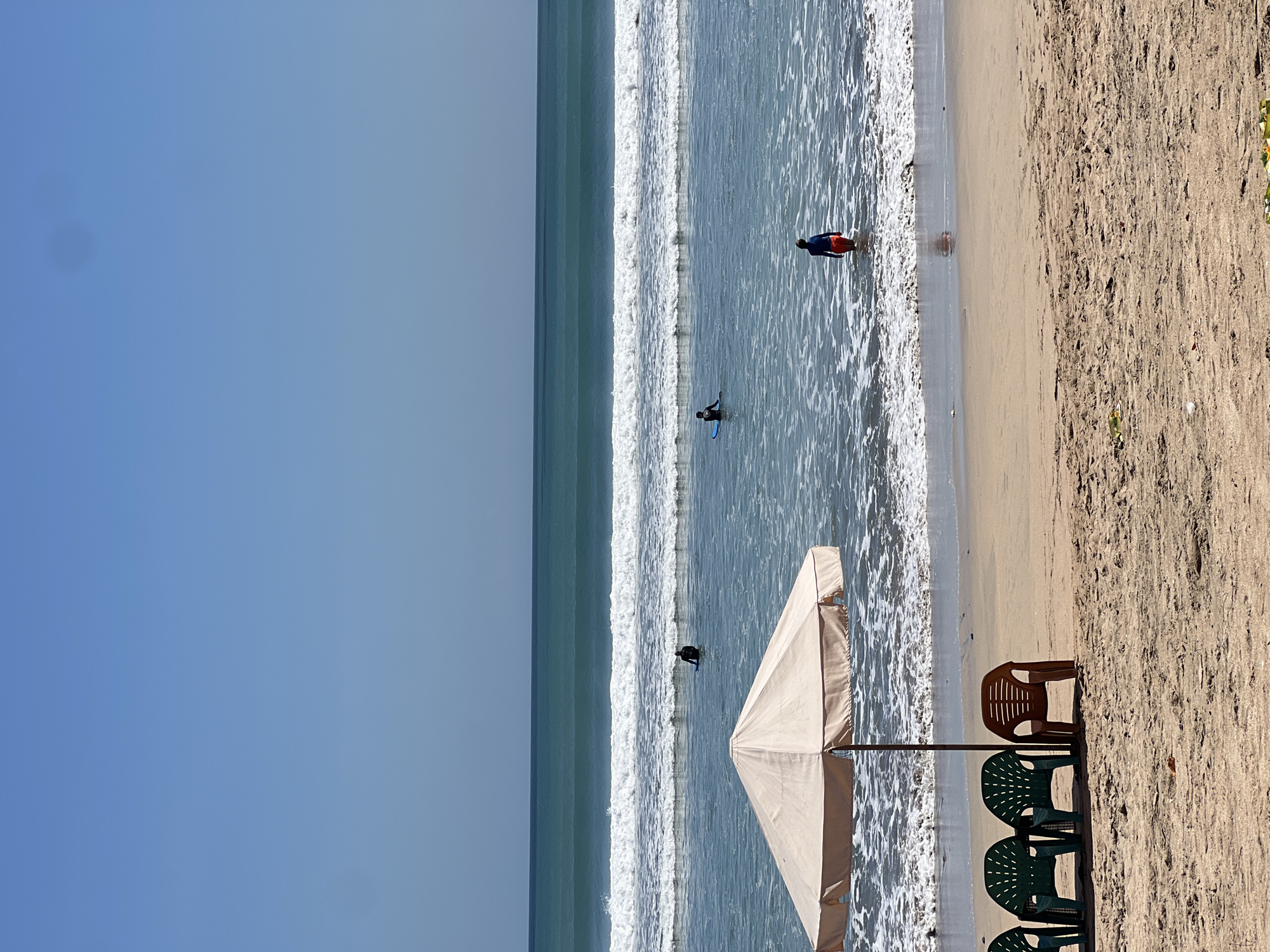 The image captures a tranquil beach scene in Kuta, Indonesia, under a clear blue sky. In the foreground, several empty plastic chairs sit on the sandy shore, shaded by a large, light-colored umbrella. The ocean stretches out in the background with gentle waves rolling in. Several individuals are visible in the water: one person is sitting on a surfboard, another is wading, and a third is further out, appearing to surf. The environment is a typical beach setting, suggesting a warm, sunny day.