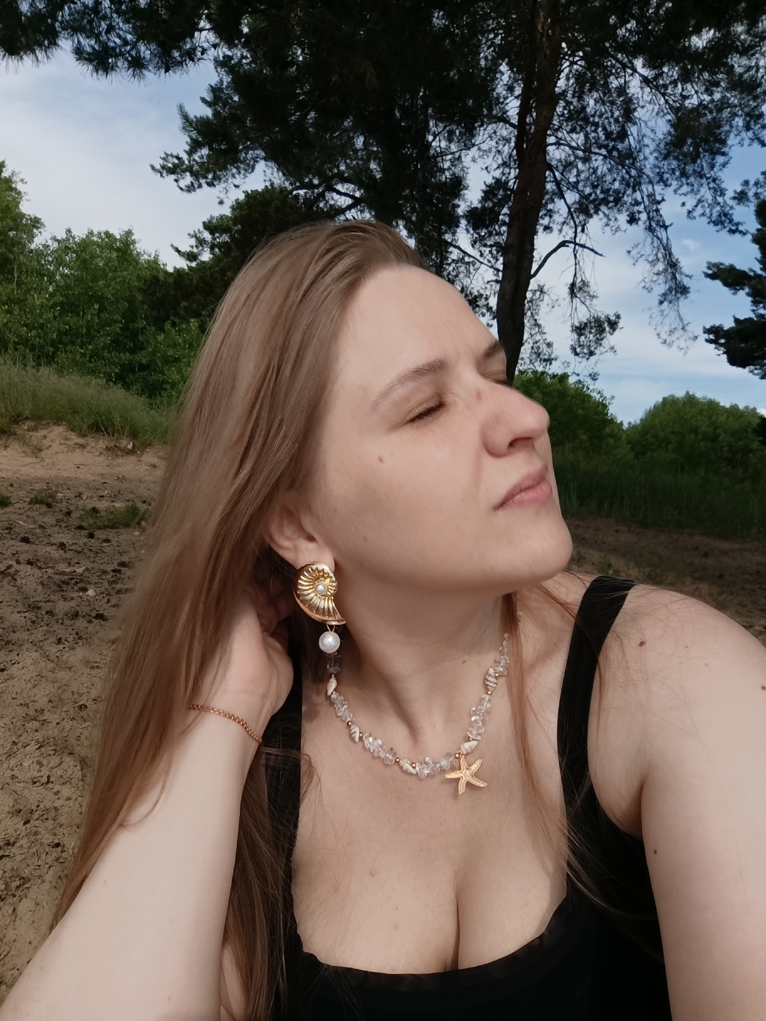 A close-up, medium shot shows a Caucasian woman with long blonde hair and fair skin, wearing a black tank top, a gold seashell earring, and a necklace with a starfish pendant and clear beads. Her eyes are closed, and her head is tilted back slightly, as if basking in the sun. The background is a blurred natural scene with trees, sand, and a blue sky. The image was taken outdoors in an unspecified location, possibly a beach or park, given the sandy terrain and foliage.