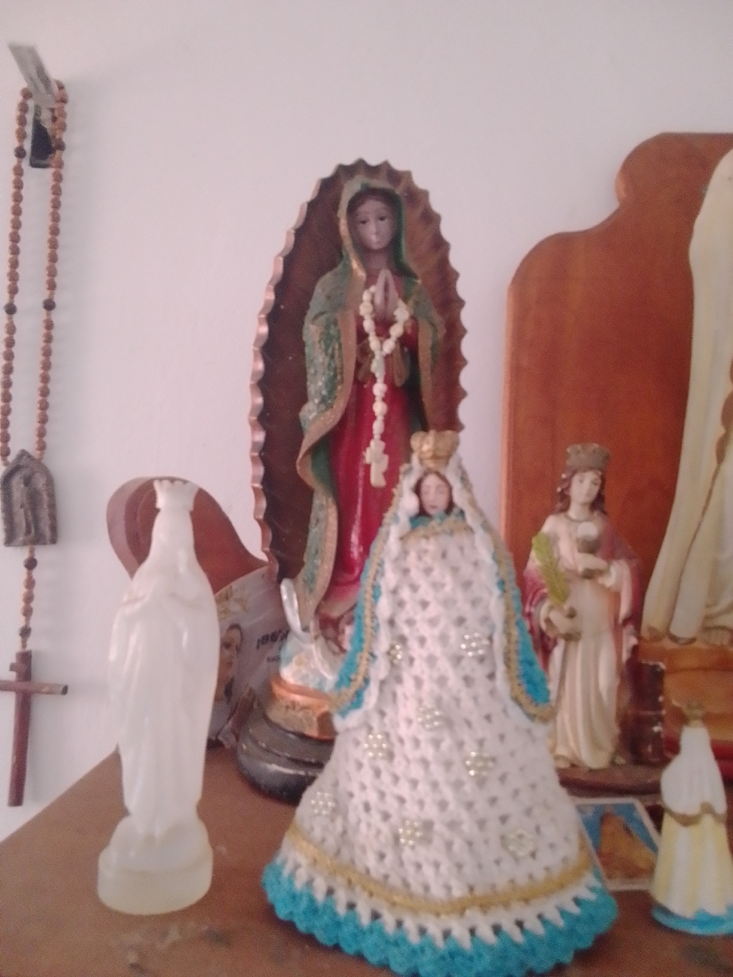 This media file is a still photograph of a religious altar. The altar is situated against a white wall, likely indoors. The primary subjects are several figurines of religious figures, most prominently the Virgin Mary.
On the left, a strand of brown rosary beads hangs from a small religious medal, with a wooden cross further down the strand. Next to the rosary, a tall, slender, white translucent figurine of the Virgin Mary stands on a shelf.
In the center of the image, a larger figurine of the Virgin Mary, depicted as Our Lady of Guadalupe, is displayed within an ornate brown arch. She is adorned in traditional red and green robes and a white rosary.
In front of this central figure, a richly decorated figurine of the Virgin Mary is prominently displayed. She wears a crown, and her gown is a white, crocheted overlay adorned with small, pearl-like embellishments and trimmed with gold and blue.
To the right of the central Virgin Mary, another figurine of a female saint, possibly holding a palm frond, stands. Further to the right, a very small, simple figurine with a yellow and blue base is visible. The altar itself appears to be a wooden shelf.
The lighting suggests an indoor setting, possibly during daylight hours, with the light source coming from the front, casting subtle shadows. There are no clear indicators of the specific time of day or weather conditions. The overall mood is one of reverence and devotion.
No visible text is present in the image, except for what appears to be a very small portion of text on a picture behind the white figurine on the left.
The context provided is "La Tagua, Venezuela," indicating the likely geographical location of this religious display.