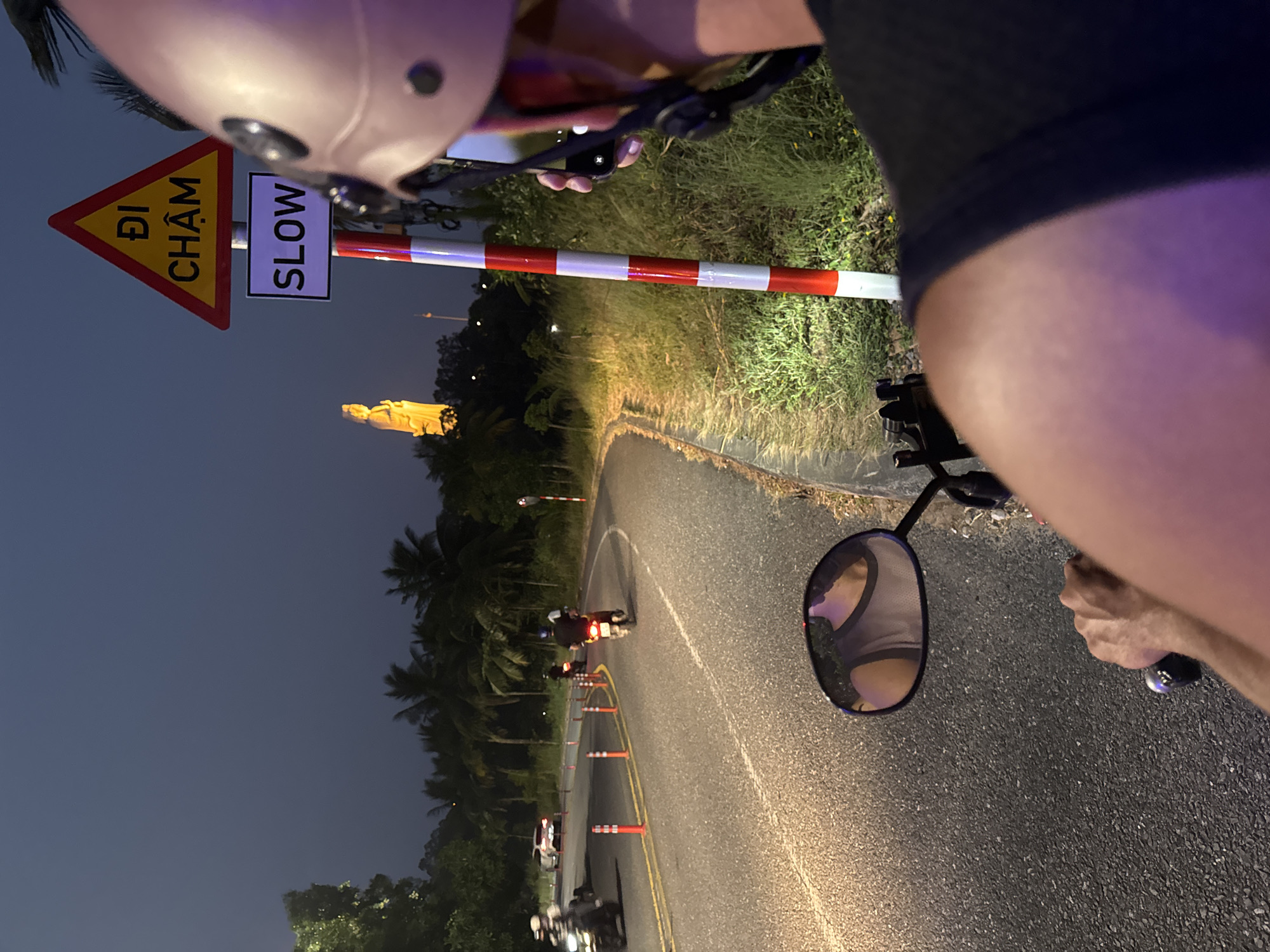 The photograph, taken from the perspective of a person riding a scooter at night, depicts a road scene in Đà Nẵng, Vietnam. The road curves, marked by orange traffic cones and a yellow line. Several scooters are visible ahead, and a red-and-white striped traffic barrier spans the road. A sign reads "ĐI CHẬM" (Vietnamese for "GO SLOW") and "SLOW" in English. In the distance, a large, golden statue is illuminated. The scooter rider is wearing a helmet, and their arm and hand are visible. The environment includes trees and vegetation lining the road. The sky is dark.