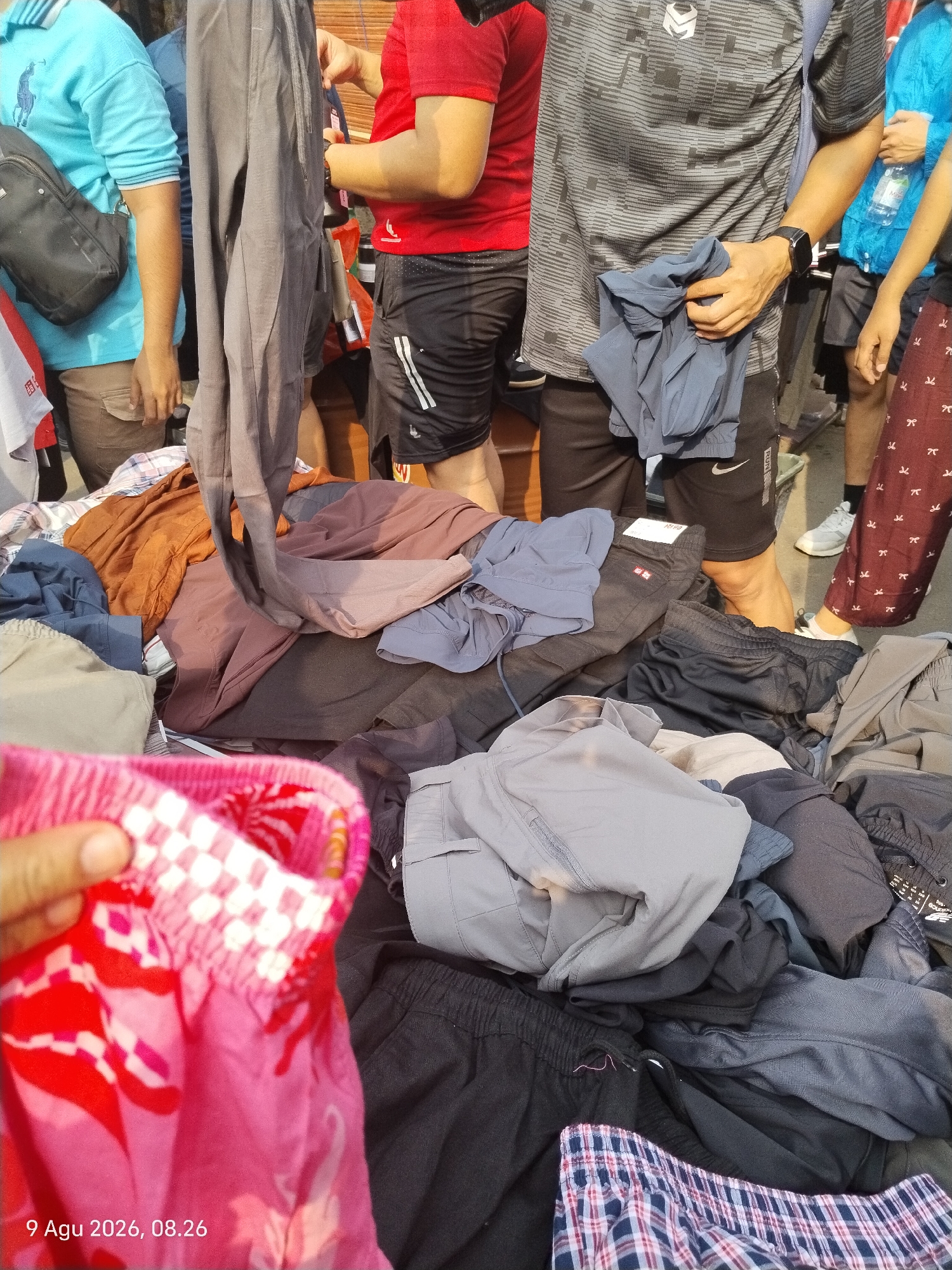 A close-up, eye-level shot captures a chaotic outdoor market scene, likely in Central Jakarta, Indonesia, focusing on a pile of folded and draped garments, predominantly pants and shorts, displayed for sale. The lighting suggests it is daytime. The foreground is dominated by a hand reaching out, holding a pair of bright pink and red patterned shorts with a white checkered elastic waistband. Behind this, a substantial assortment of casual wear is visible. These include dark gray and black cargo shorts, various shades of blue and gray athletic shorts, and several pairs of dark trousers, some with visible tags. The fabric textures range from smooth synthetic materials to textured cotton blends.
In the mid-ground and background, several individuals are partially visible, their presence indicating they are likely vendors or shoppers. On the far left, a person wearing a light blue collared shirt with a dark crossbody bag is seen. Adjacent to them, a person in a red t-shirt is visible. Further into the scene, a person in a gray and black patterned t-shirt with a black digital watch on their left wrist is holding a folded pair of dark blue shorts. Another individual on the right is wearing burgundy pants with a white pattern. The background is indistinct, suggesting the market is situated along a street or in an open area. The timestamp "9 Agu 2026, 08.26" is superimposed in the lower left corner.