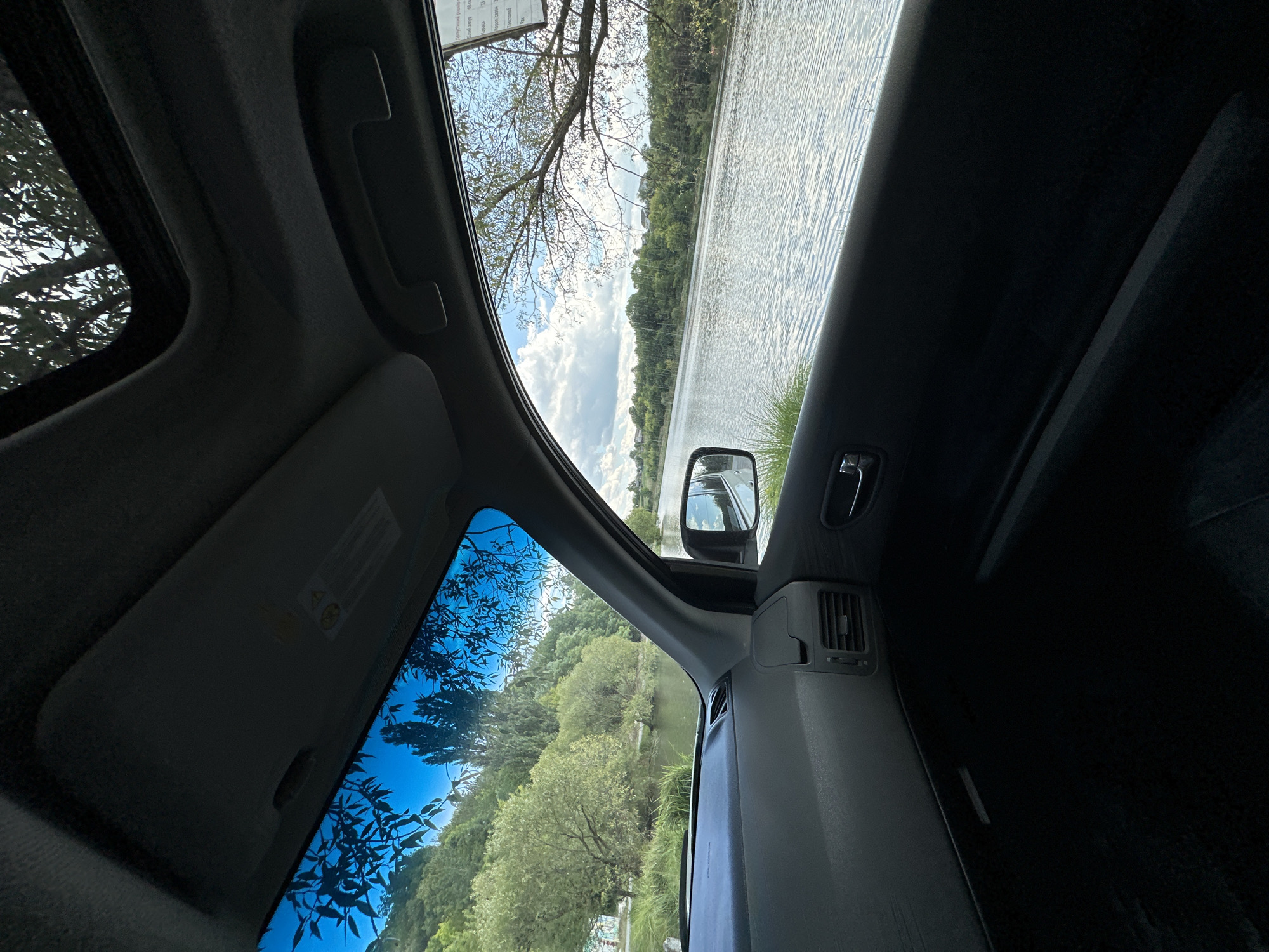 The image shows a view from the inside of a car looking out a window towards a body of water and a tree-lined shore. The car interior, including the dashboard, sun visor, and part of the door panel, is visible in the foreground. The exterior scene depicts a lake or river with rippling water, reflecting the cloudy sky. On the far bank of the water, there are trees and some houses. The sun visor is partially open, and a sunroof is visible above. A side mirror shows a reflection of the car and the environment. The scene suggests a location near a body of water, possibly a recreational area, in or near Makiv, Ukraine.