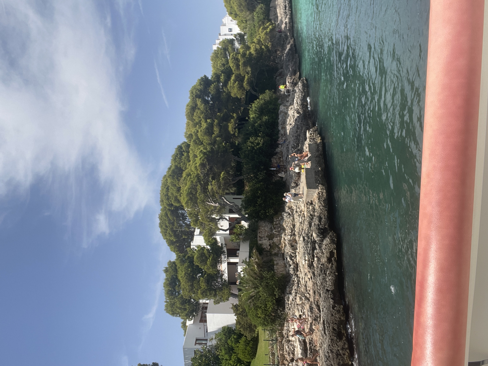 The image shows a sunlit coastal scene in Cala D'or, Spain. White buildings with flat roofs are nestled among lush green trees on a rocky hillside overlooking a turquoise sea. Several people are visible on the rocks near the water, some appearing to be sunbathing or relaxing. A concrete jetty extends into the water, with a few individuals standing on it. The water itself is clear and a vibrant blue-green, with some small waves lapping against the shore. The sky above is a bright blue with scattered white clouds. A red railing is visible in the foreground, partially obscuring the view of the water.