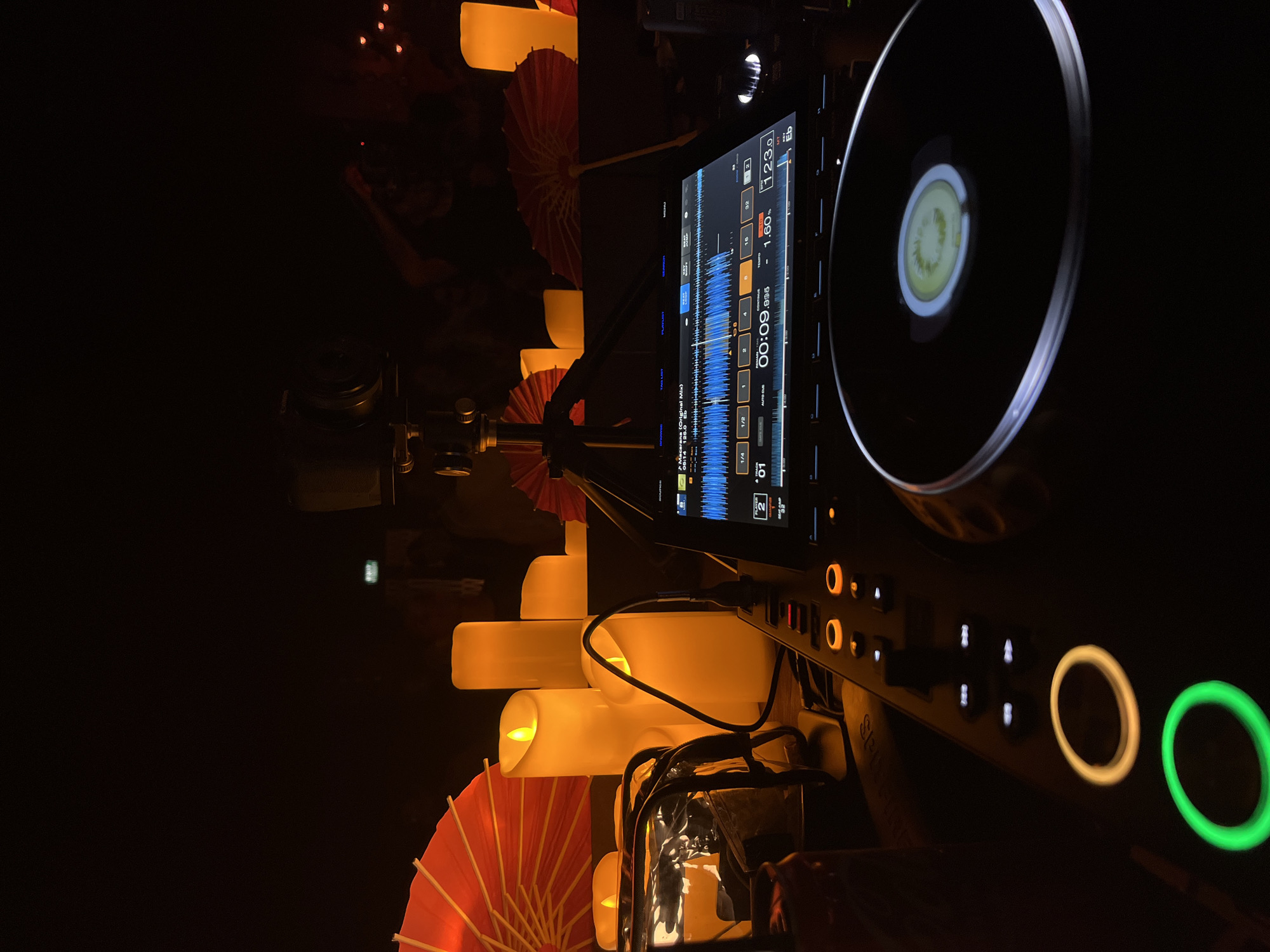 The image depicts a dimly lit, atmospheric scene, likely at a night event or performance, with a DJ setup in the foreground. Several large, illuminated LED candles of varying heights stand prominently, casting a warm glow. To the left, a clear plastic container with some items inside is visible. In the background, several red paper parasols are arranged, adding a touch of decorative color to the dark environment. A DJ mixer with a screen displaying waveforms and controls is central to the foreground, with illuminated buttons and a spinning platter. The overall ambiance suggests a relaxed, possibly musical, gathering. The location in Ban Bang Thao, Thailand, is not visually evident from the image itself. There are no discernable people or direct interactions captured in the frame.